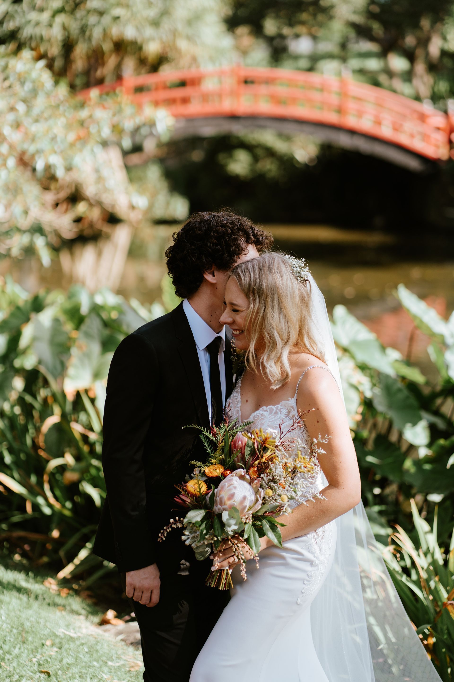 Bride and groom embrace in front of a red bridge and lake, she holds a bouquet.