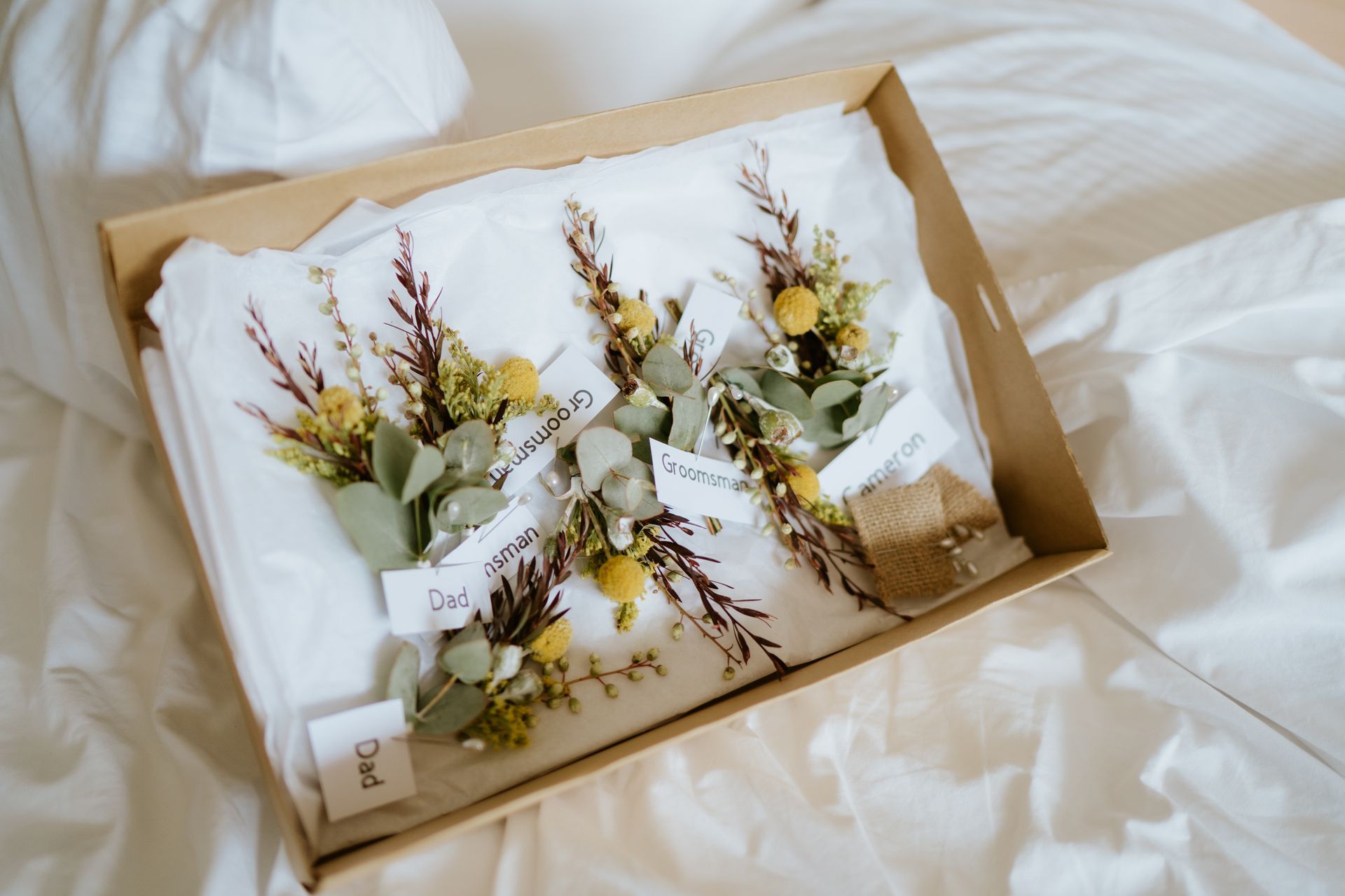 Boutonnieres in a box. Greenery, yellow flowers, name tags on a white cloth in a cardboard box.