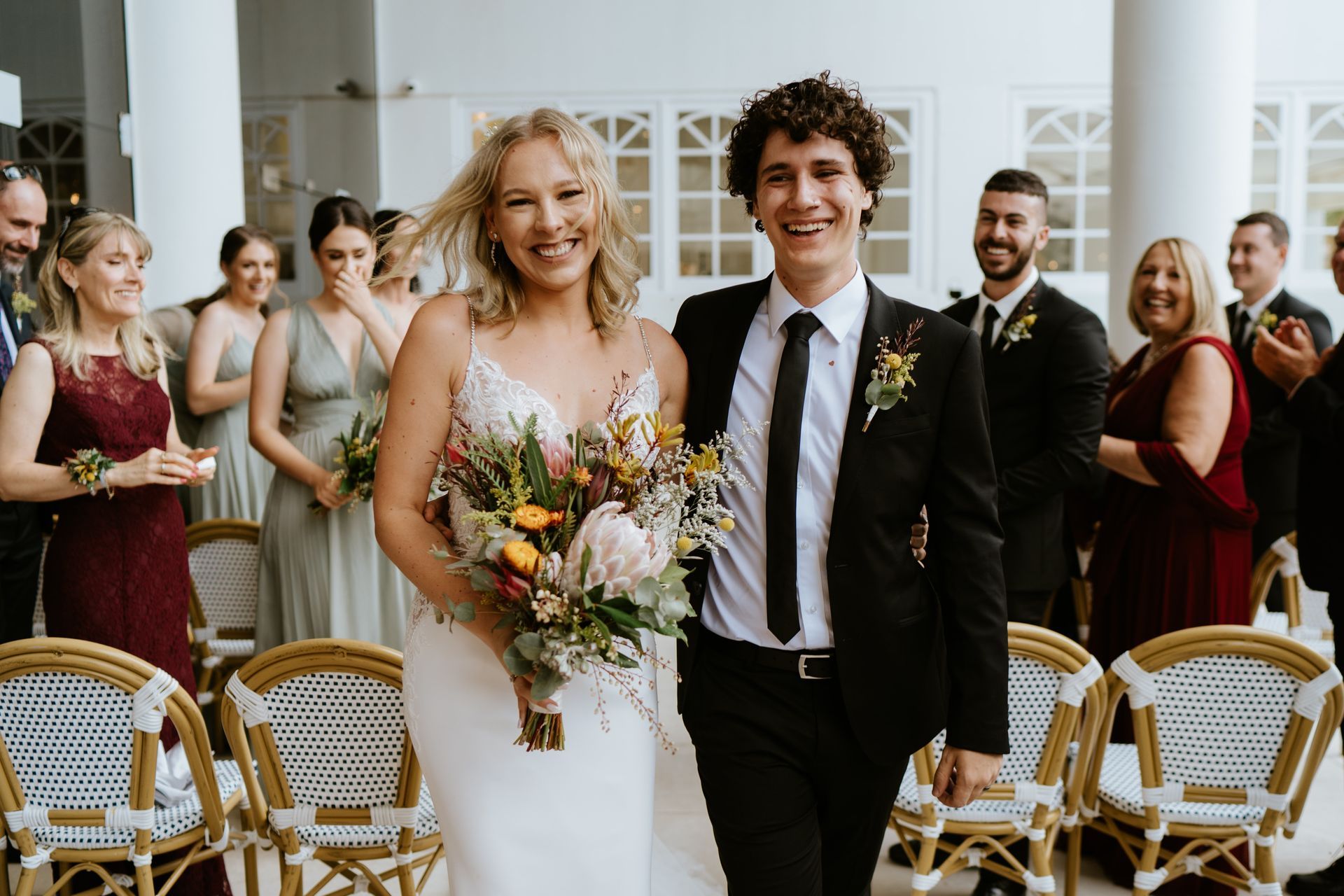 Newlyweds exiting ceremony, smiling, holding flowers. Guests clapping in outdoor setting.