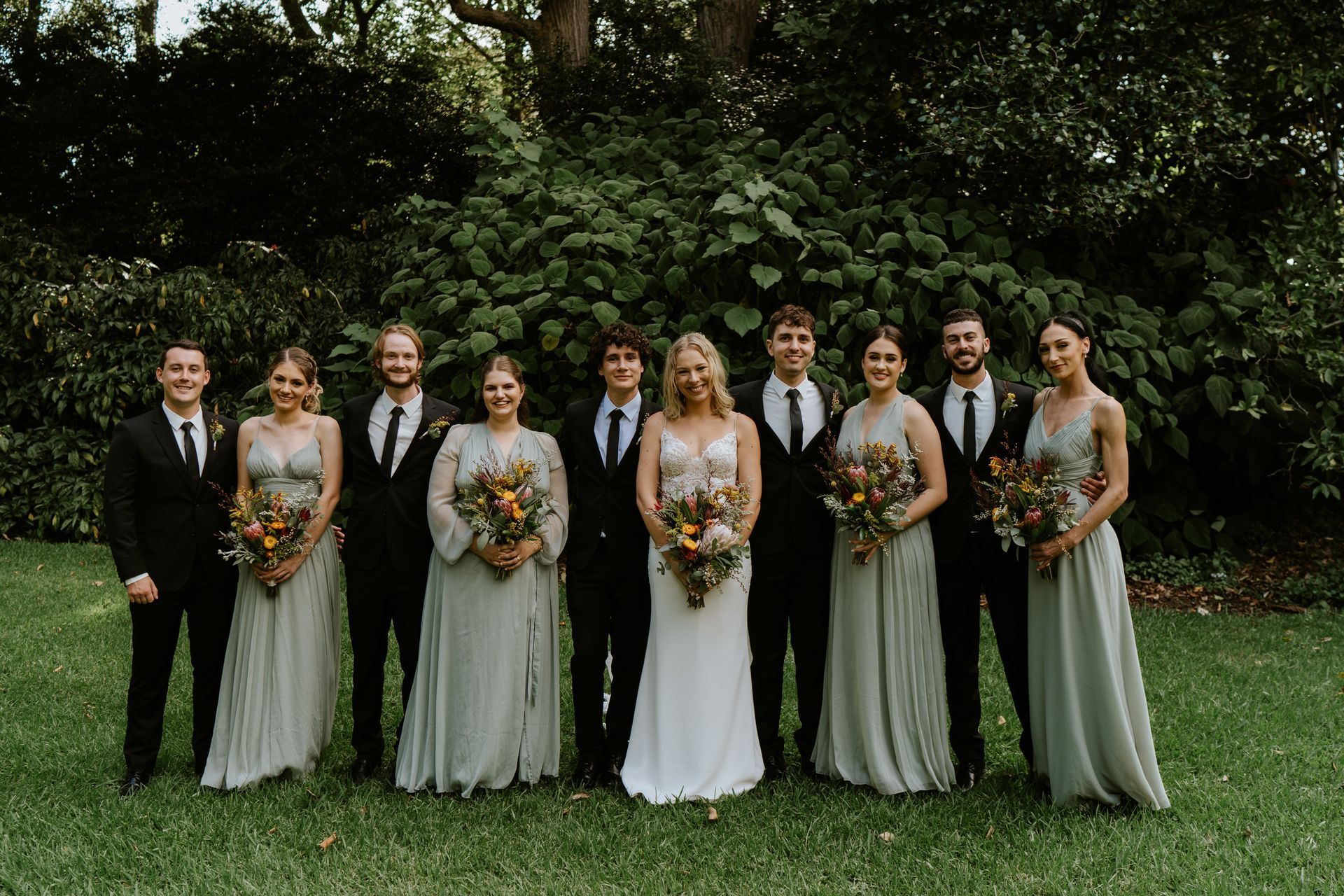 Wedding party posing in a garden. Bride in white dress, bridesmaids in green, groomsmen in black suits.