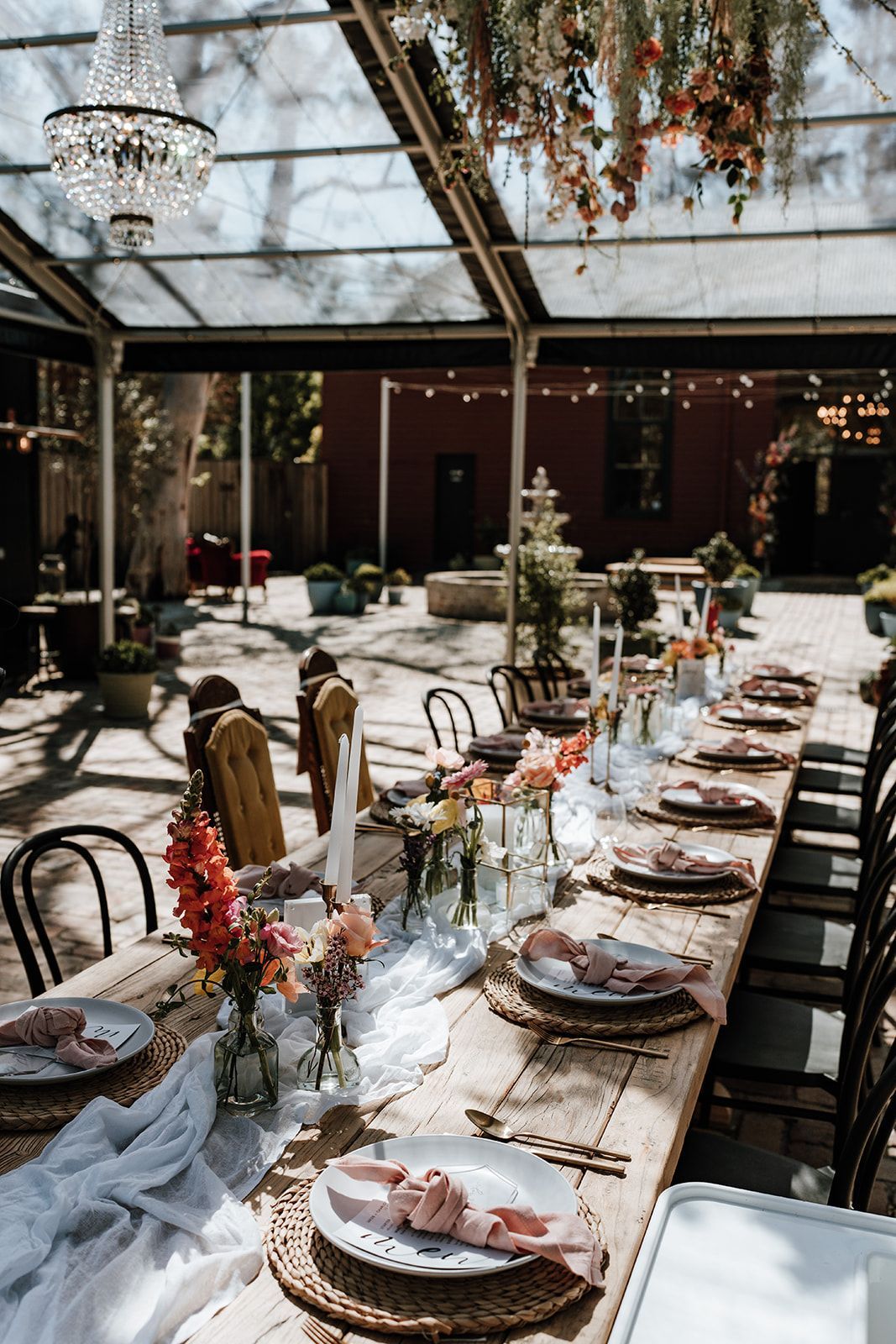 Long wooden table set for a wedding reception under a glass roof with floral decor and a chandelier.