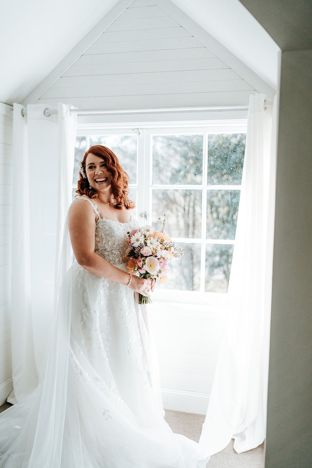 Bride with red hair in wedding dress smiles, holding bouquet by window.
