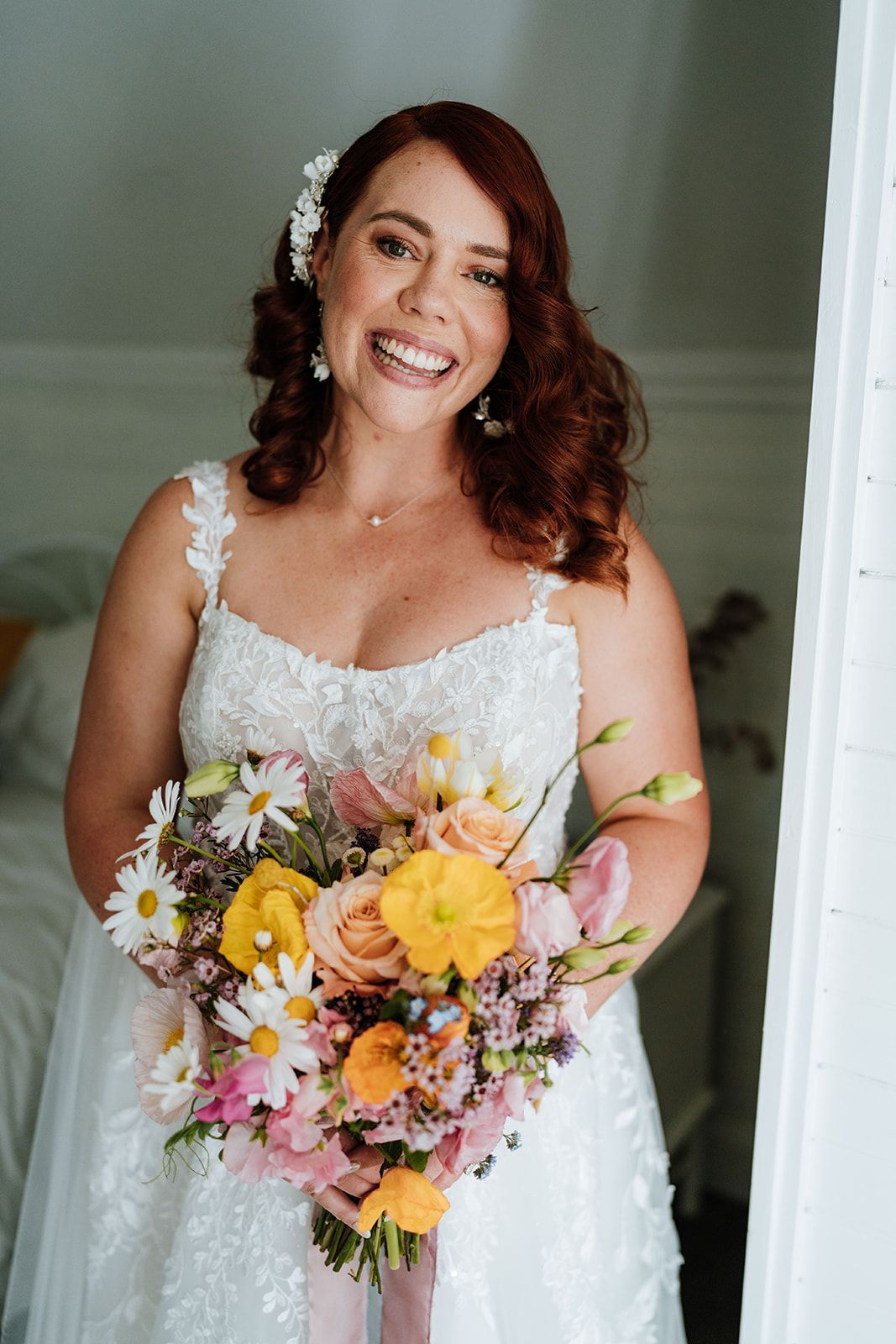 Bride in white lace gown smiles, holding colorful bouquet.  Red hair, floral headpiece. Bright, cheerful.