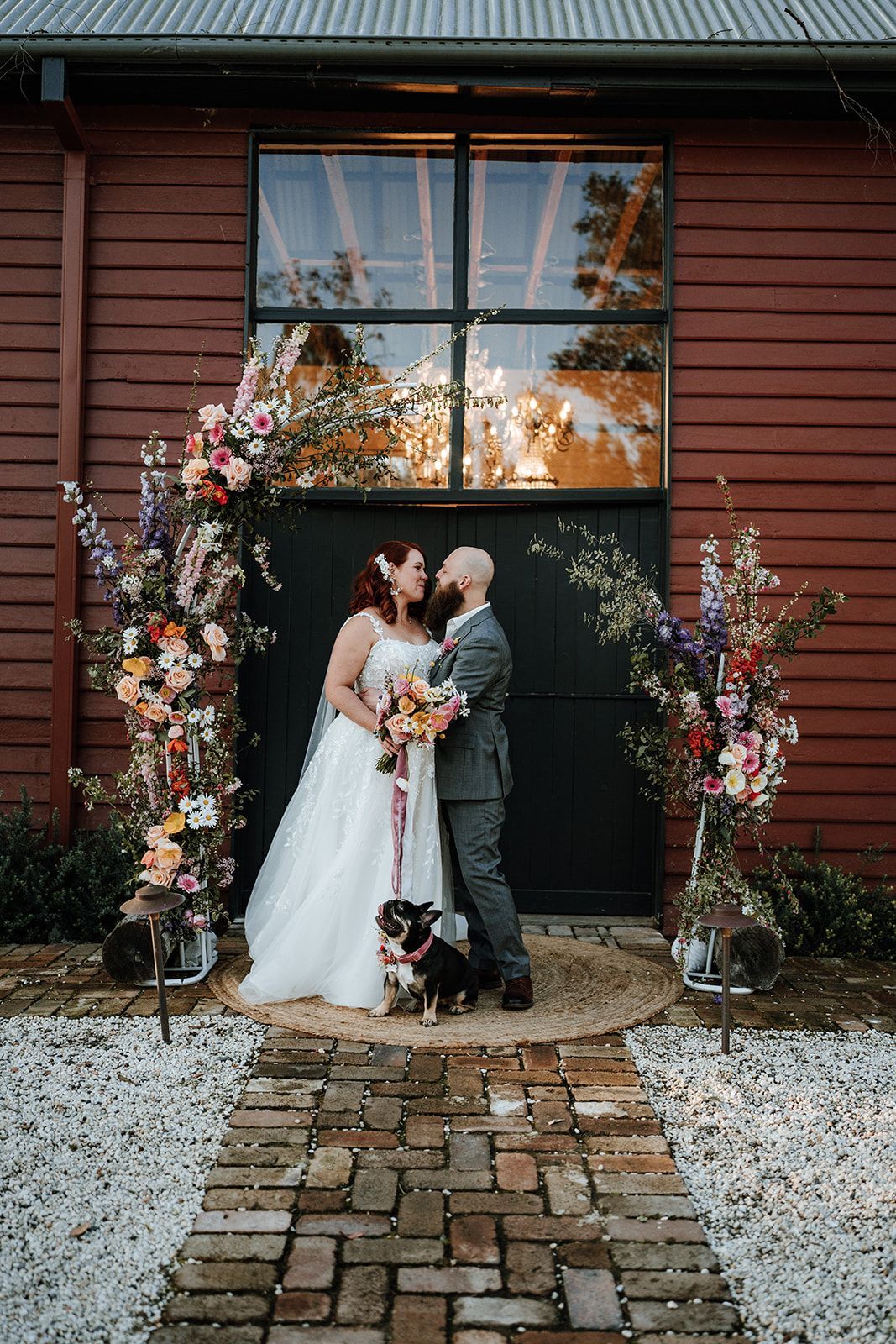 Wedding couple kissing in front of a barn doorway, framed by flowers, with a dog.