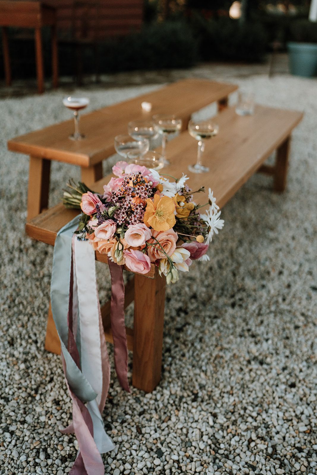 Bouquet with ribbons on wooden bench; drinks sit on the bench, gravel ground.