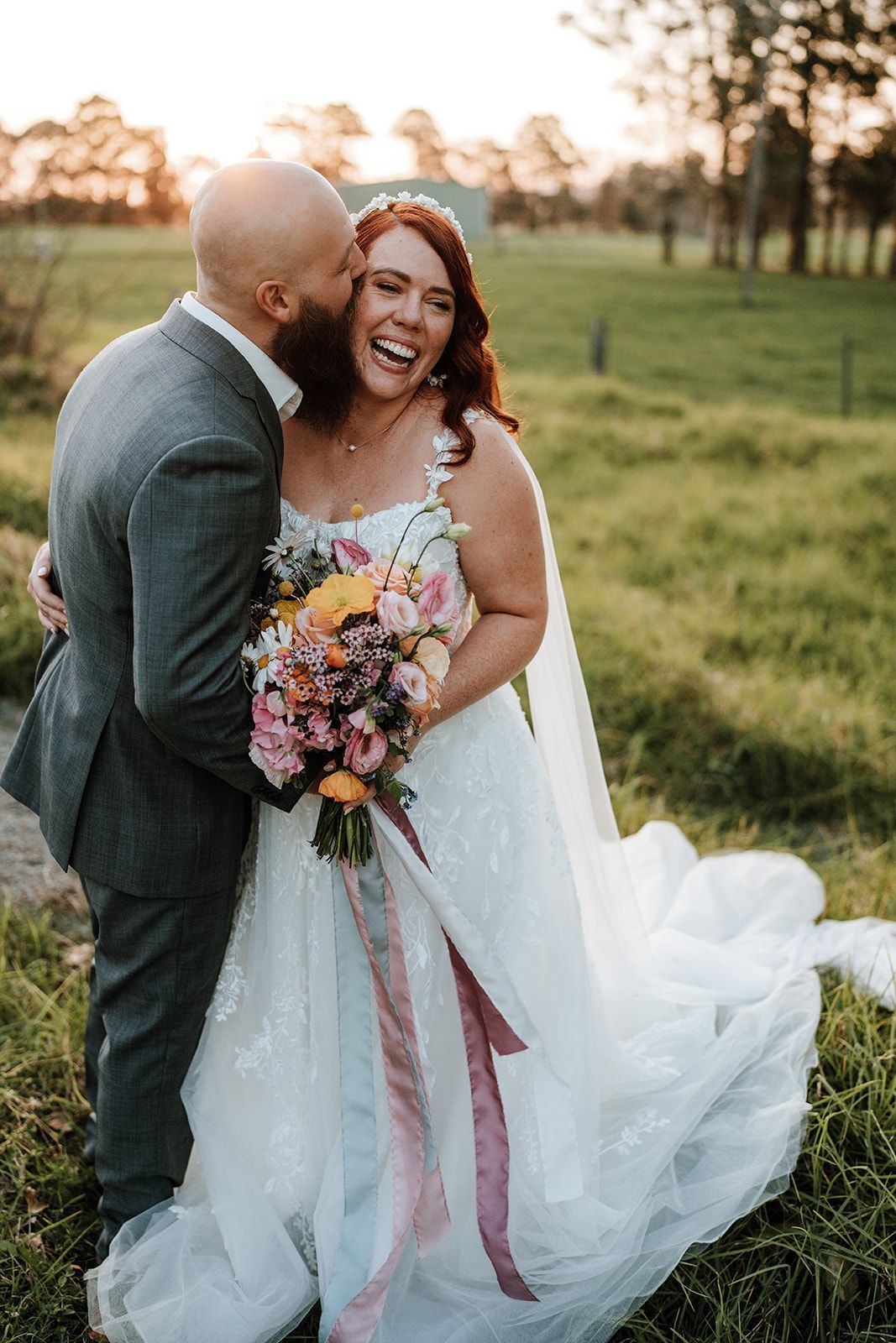 Bride and groom embrace and laugh outdoors; sunset. Bride in white gown with bouquet; groom in gray suit.