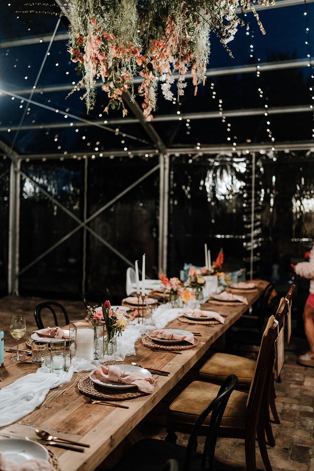 Long wooden table set for a dinner party, under a tent with fairy lights and floral decor.
