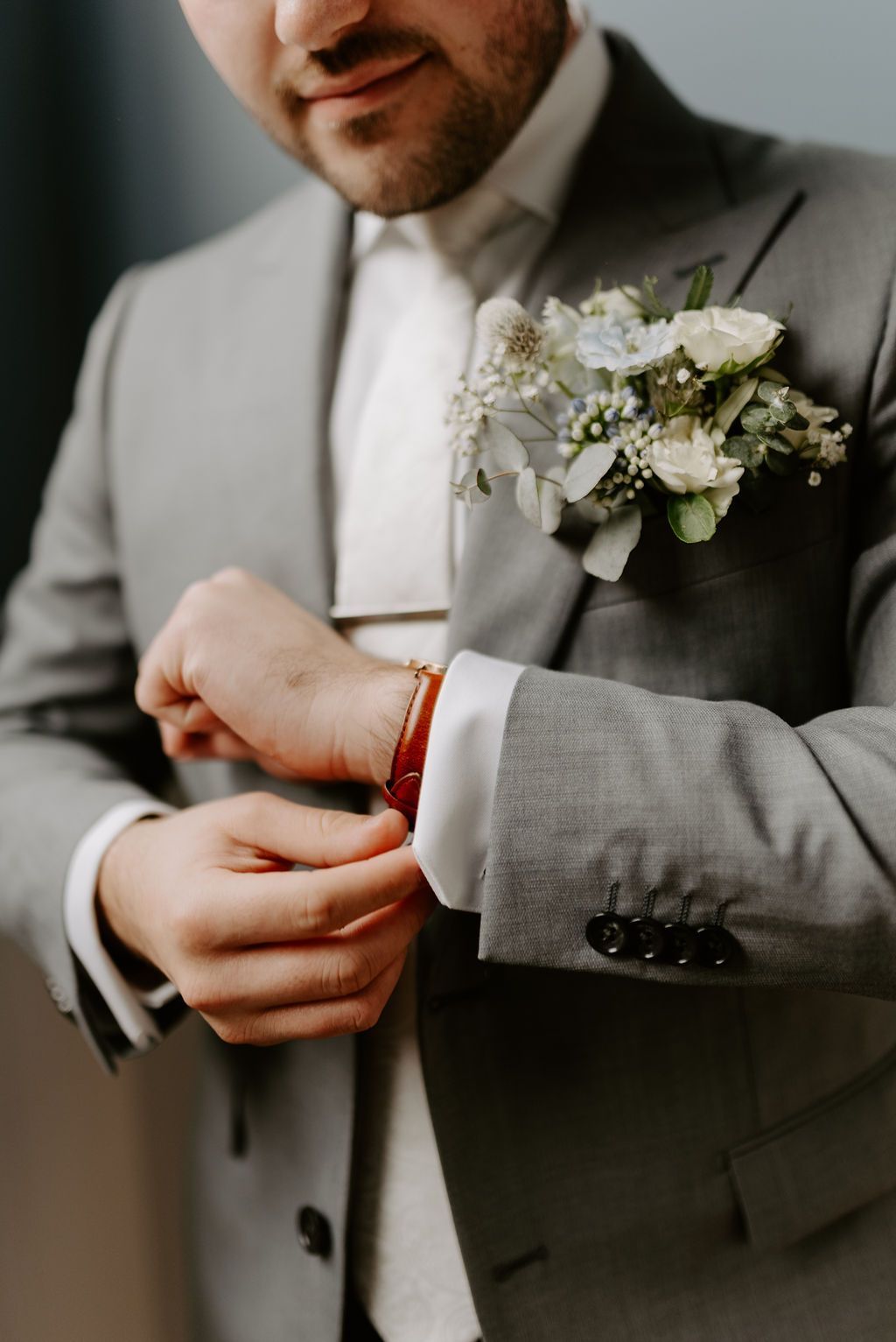 Man in a gray suit adjusting cufflink, wearing a tie and a boutonniere with flowers.