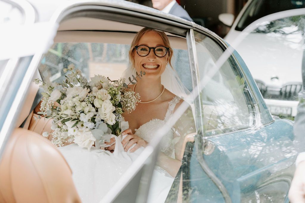 Bride in a wedding dress smiles, holding bouquet, seated in a car, looking out the window.