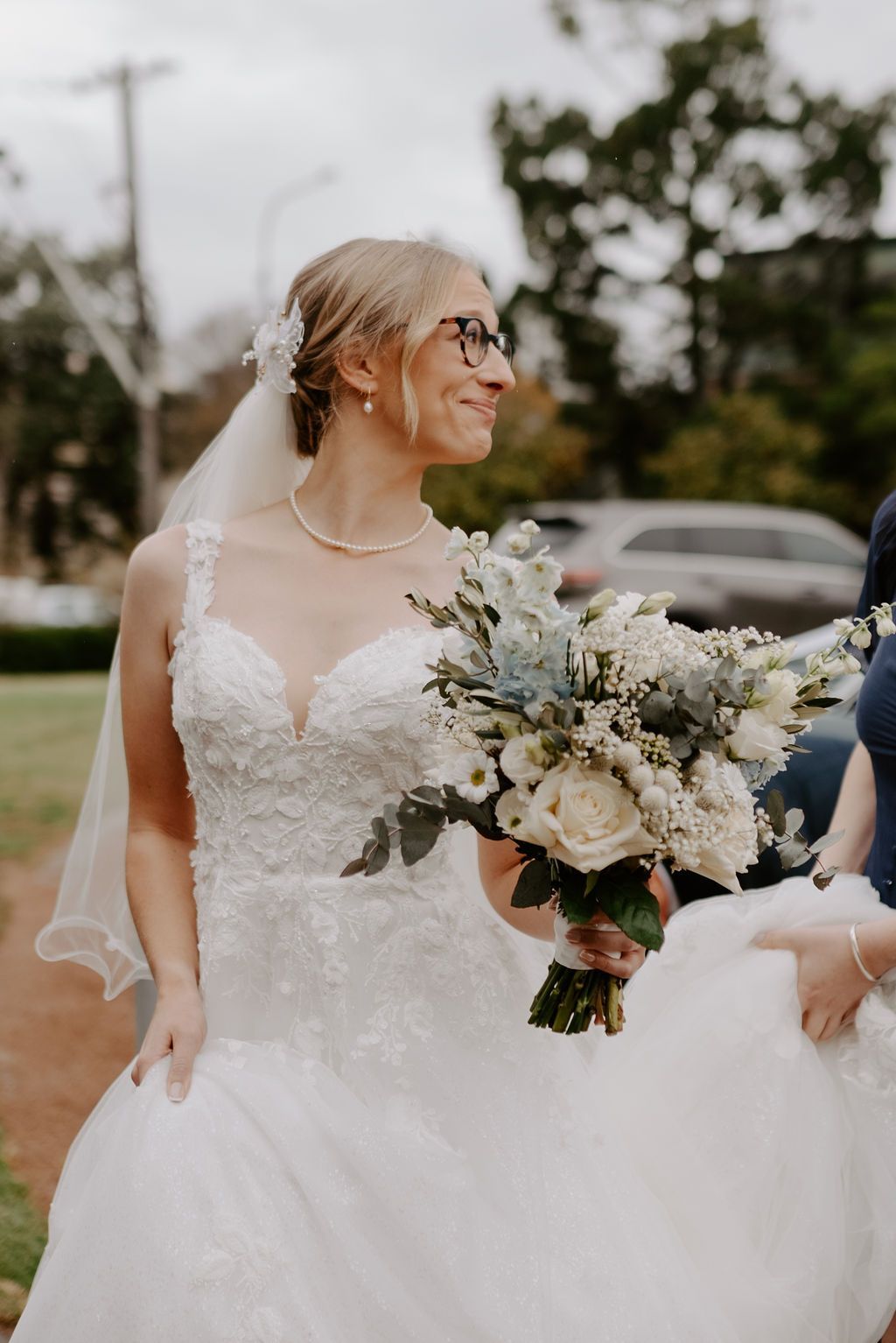 Bride in a white lace wedding dress, veil, and eyeglasses, holding bouquet, outdoors.