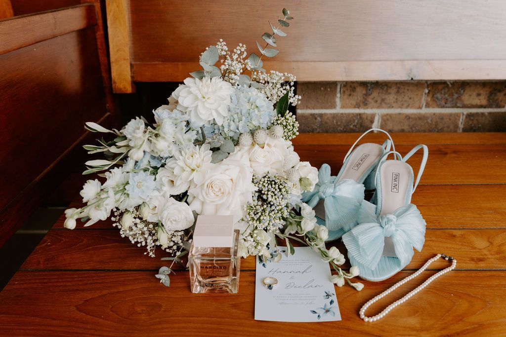 Wedding details: bouquet, light blue shoes, perfume, invitation, and necklace on wood.
