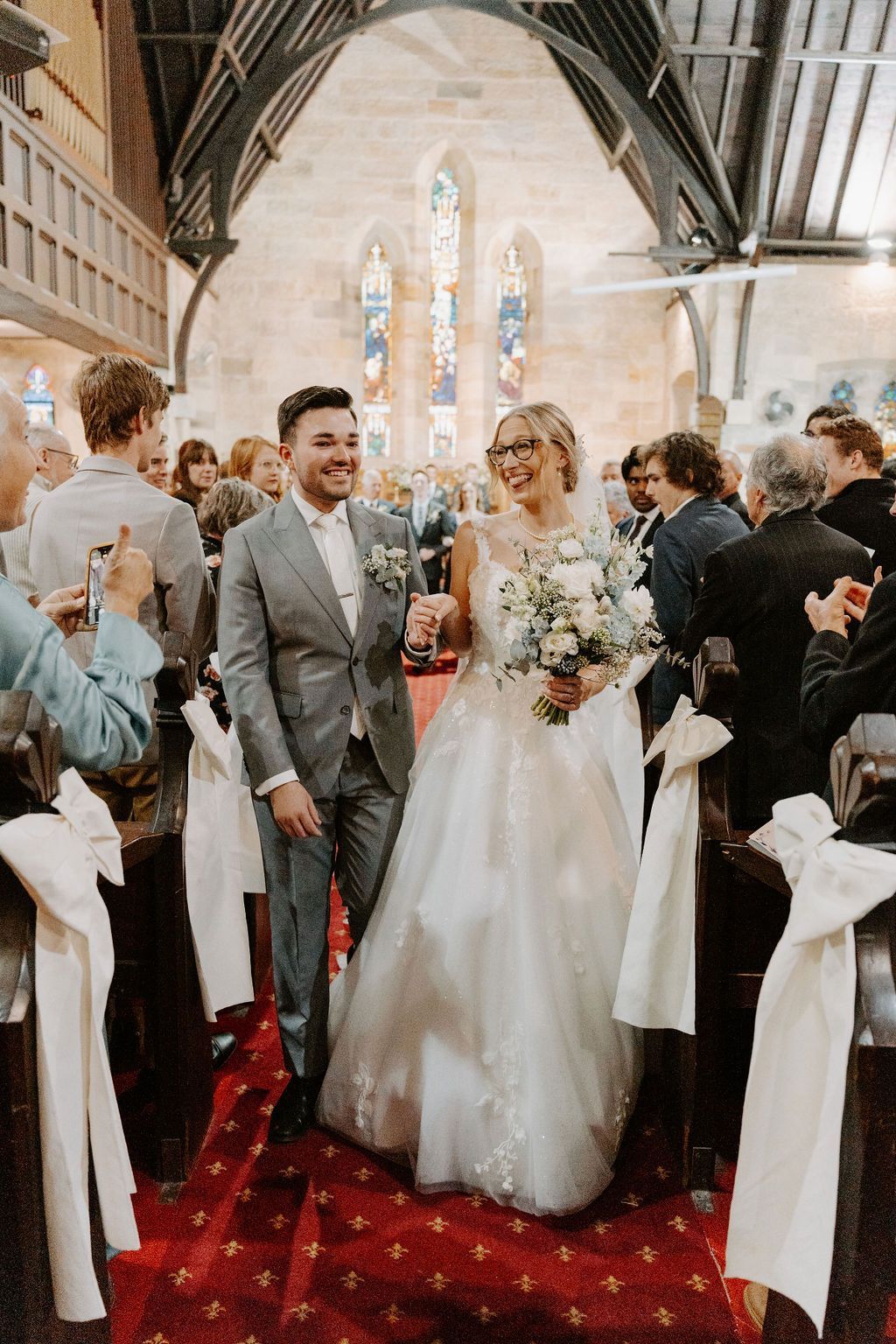 Newlyweds smiling, exiting a church after their wedding, red carpet, white bows, and guests clapping.