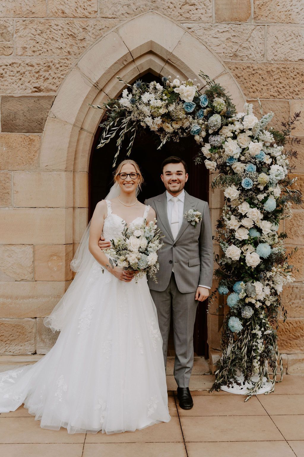 Couple in wedding attire posing under floral archway at church entrance.