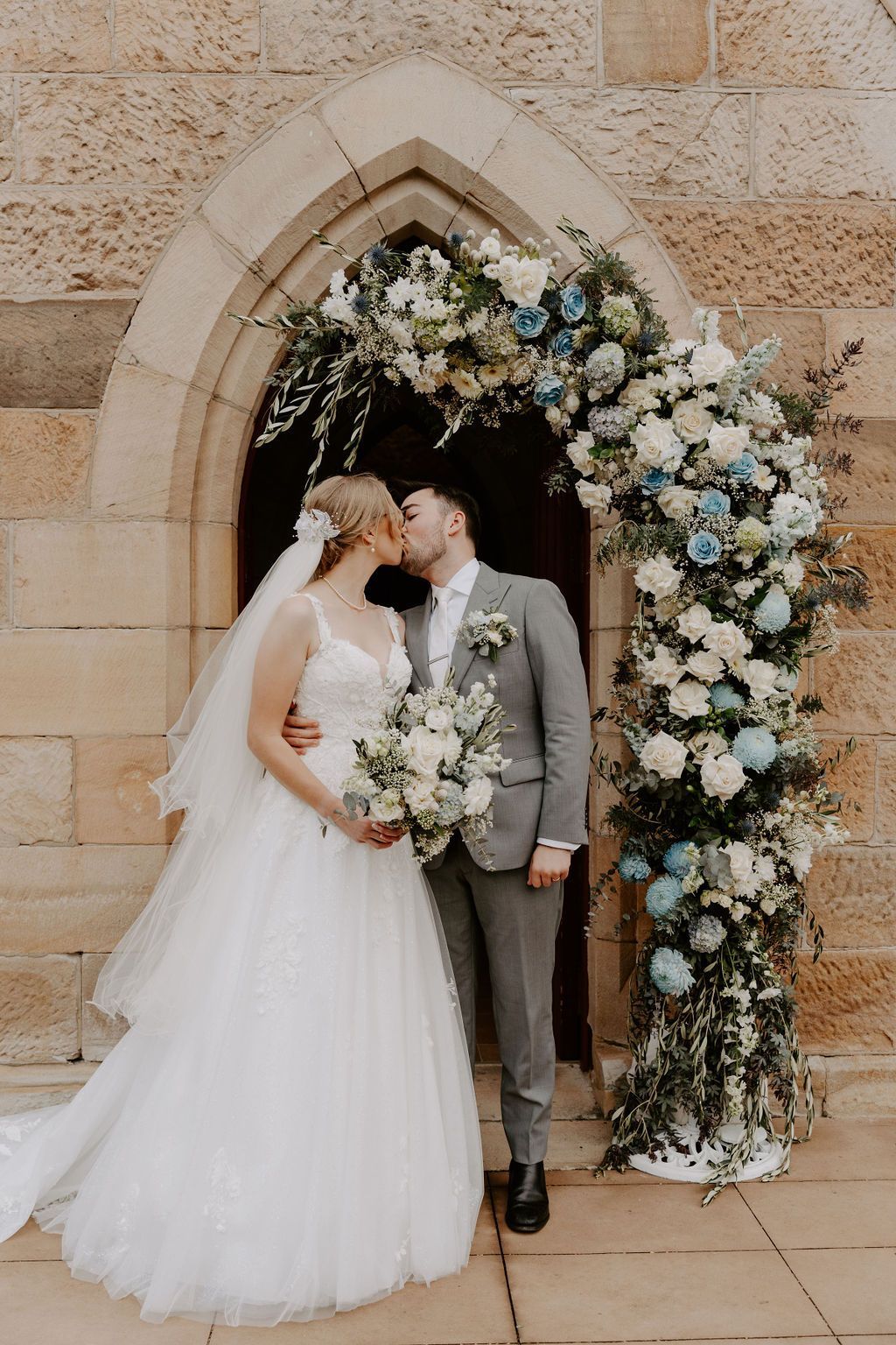 Newlyweds kissing under a floral archway at a church entrance. Bride in white dress, groom in gray suit.