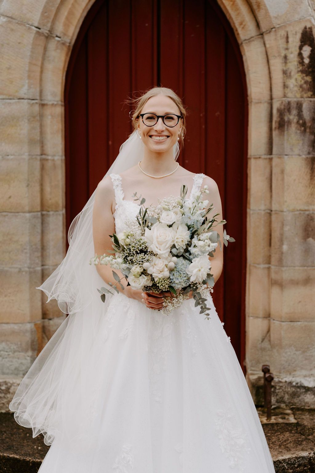 Bride in a white gown and veil holding a bouquet in front of a red door. She's wearing glasses and smiling.