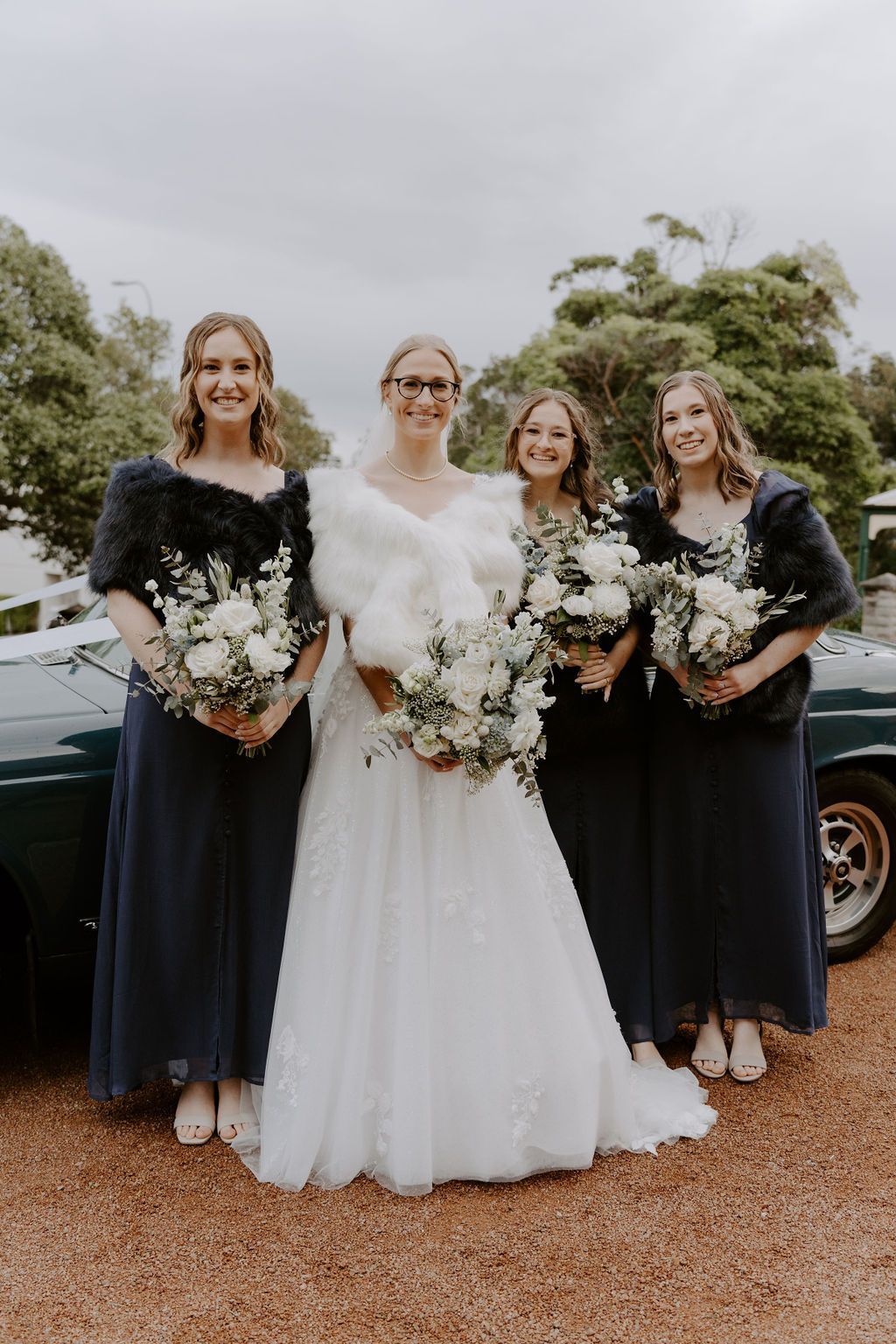 Bride with three bridesmaids in front of a green car, all holding bouquets. Bride in white, bridesmaids in navy dresses.