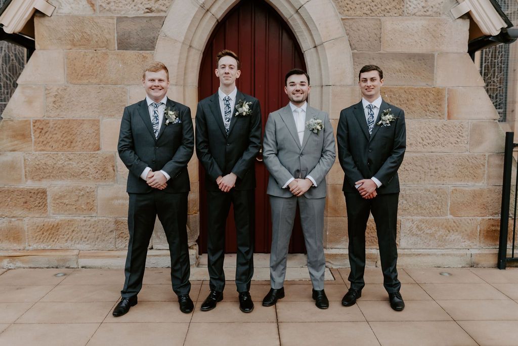 Four men in suits pose in front of a stone building with a wooden door.