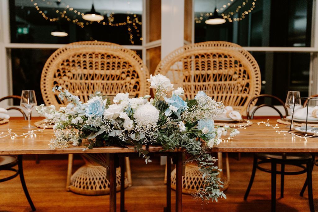 Wedding reception table setting with floral arrangement and peacock chairs.