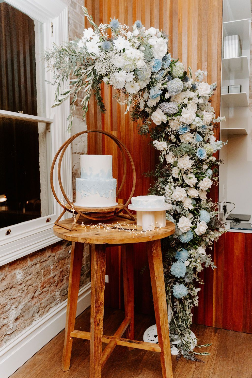 Wedding cake on a wooden table, adorned with a floral arch of white and blue flowers.