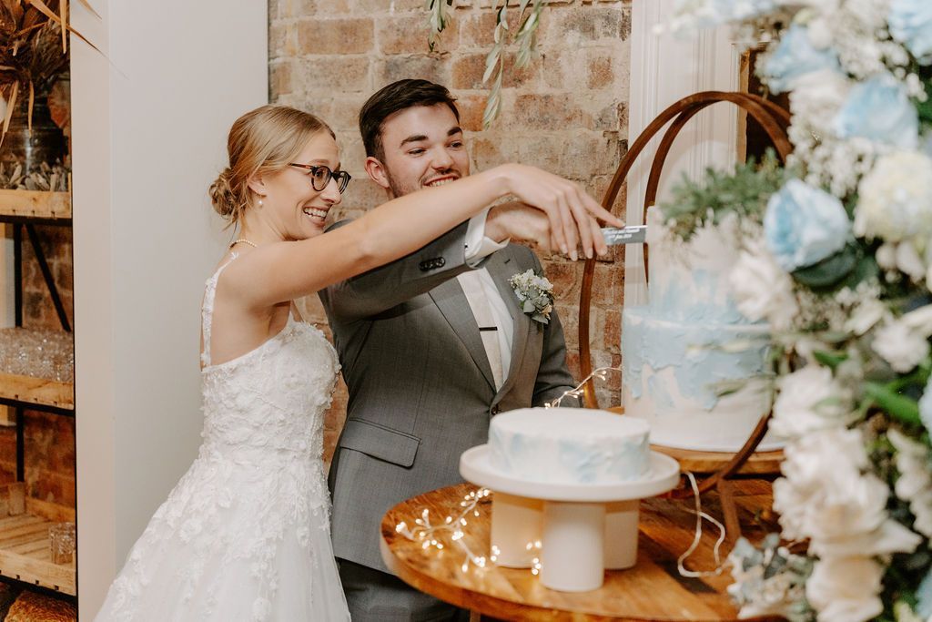 Newlyweds cutting a tiered cake at a wedding reception. The backdrop features brick and floral decorations.