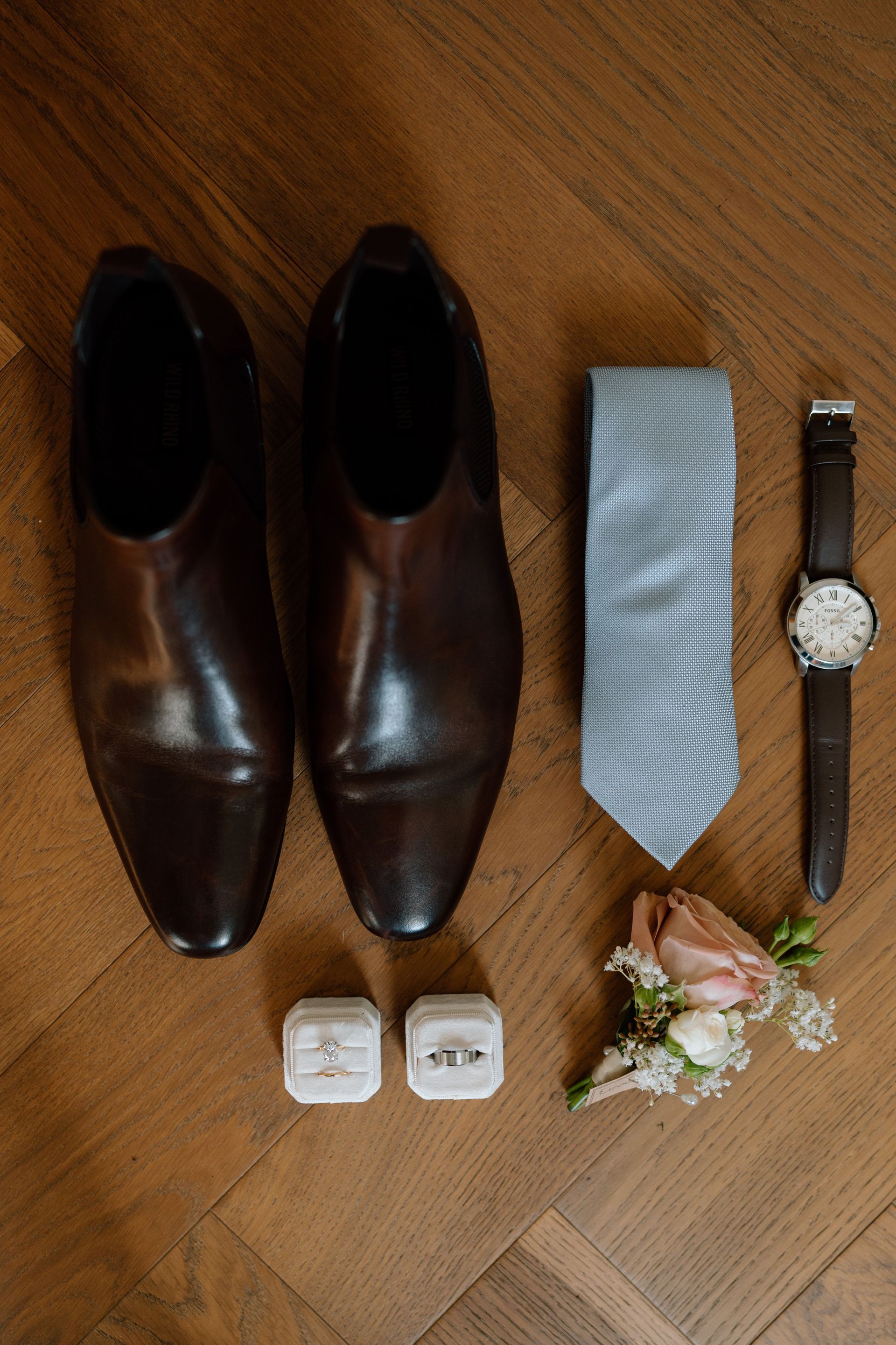 Groom's accessories: brown shoes, blue tie, watch, wedding rings in boxes, and boutonniere on a wooden floor.