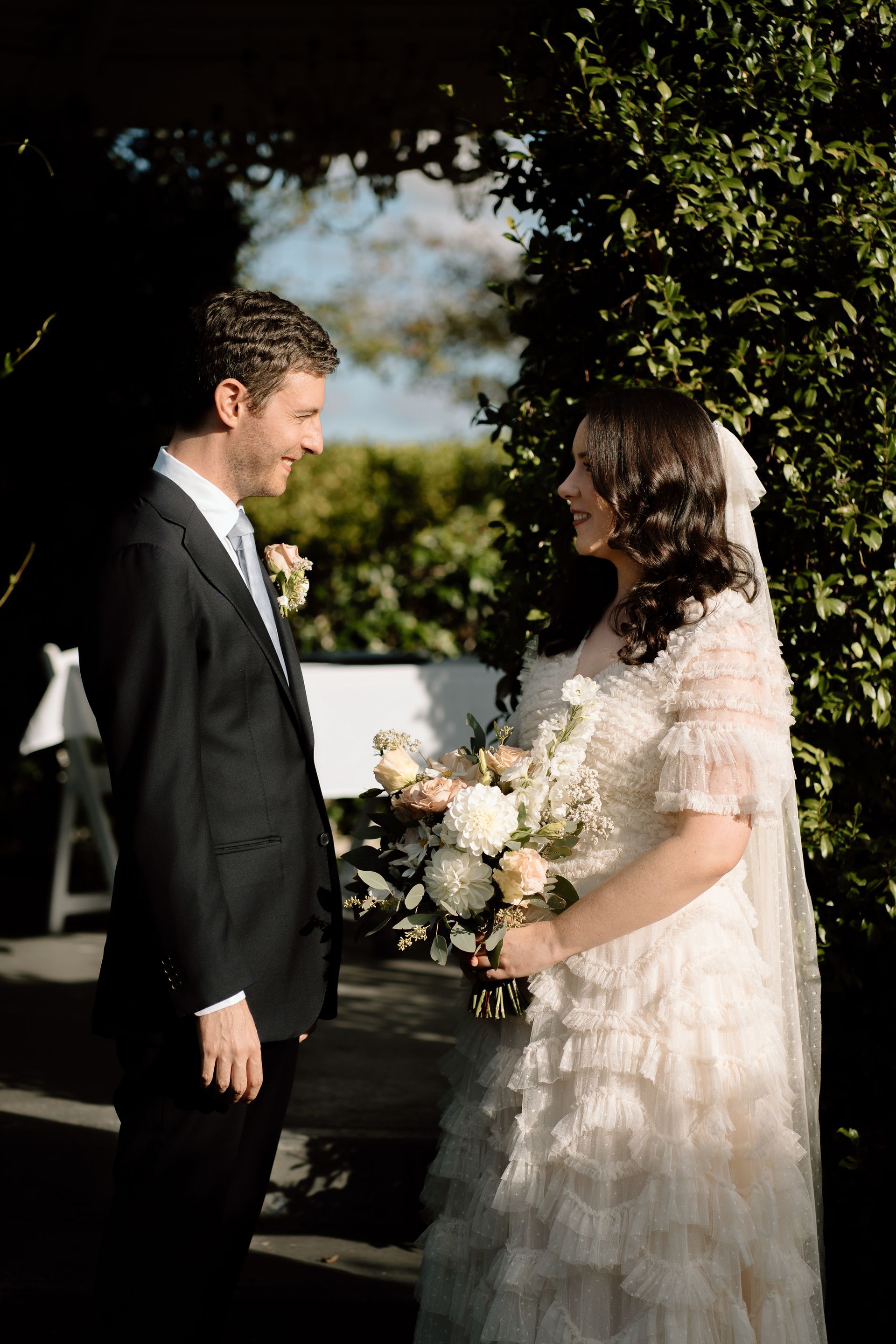 Groom smiles at bride holding flowers, both facing each other outdoors. Bride wears white dress and veil.