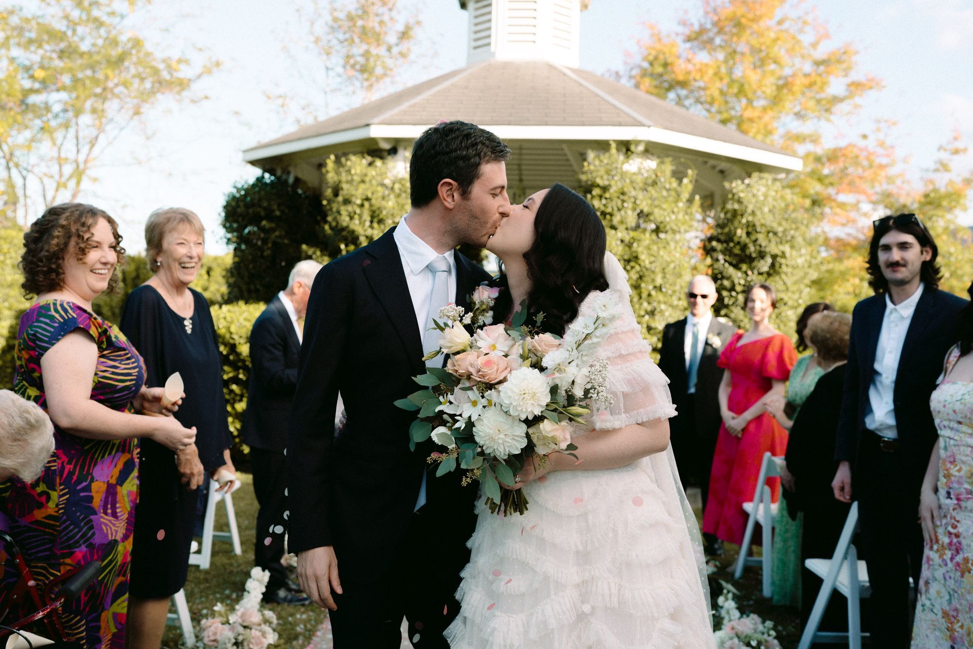 Newly married couple kissing in front of gazebo; guests in background.
