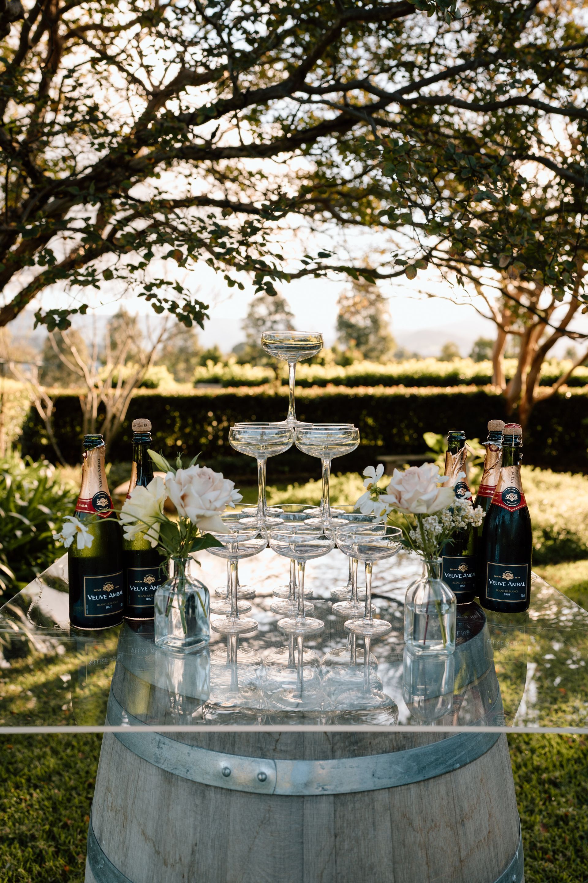 Champagne flutes on a barrel table, arranged in a tiered fountain, with bottles and flowers; outdoors.