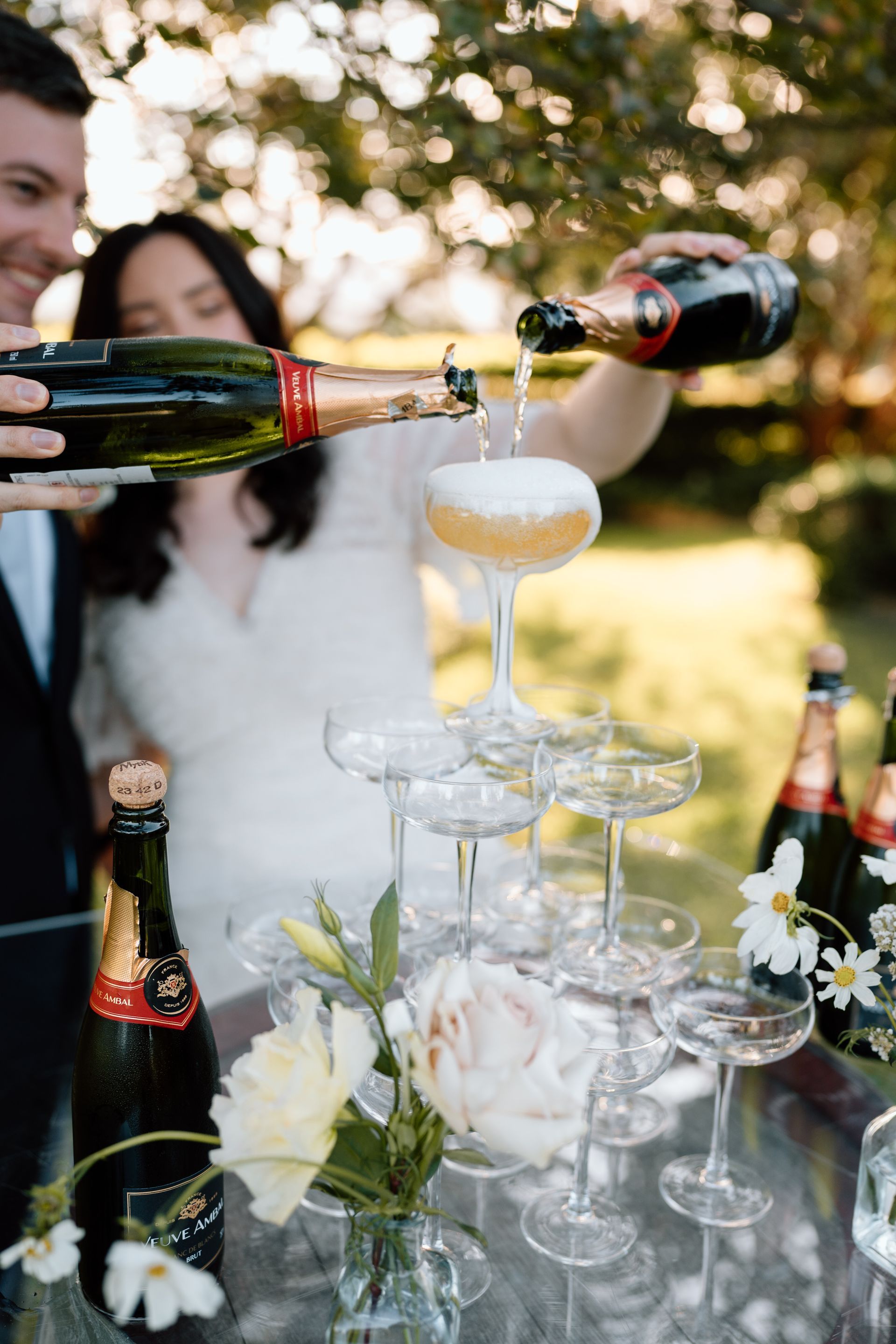Couple pouring champagne into a tiered glass tower at an outdoor wedding, with flowers and bottles visible.