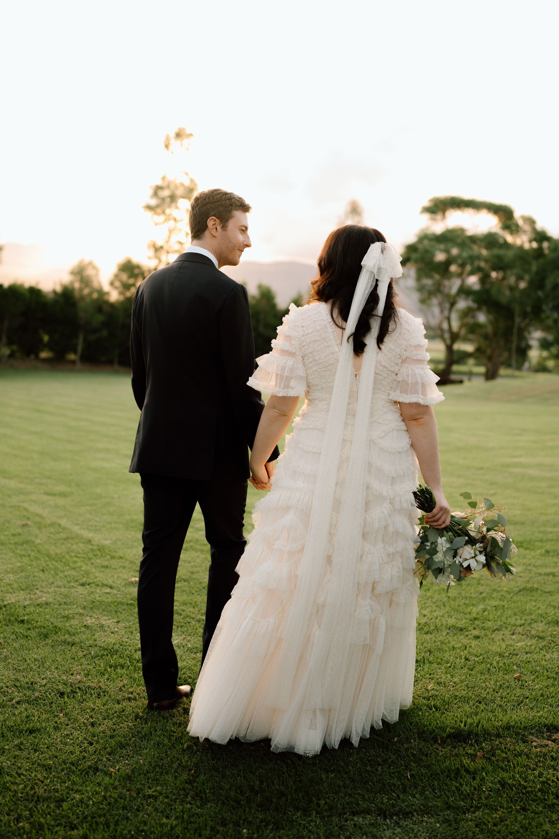 Newlyweds holding hands, walking away on a grassy field at sunset; bride in lace dress and veil, groom in a suit.