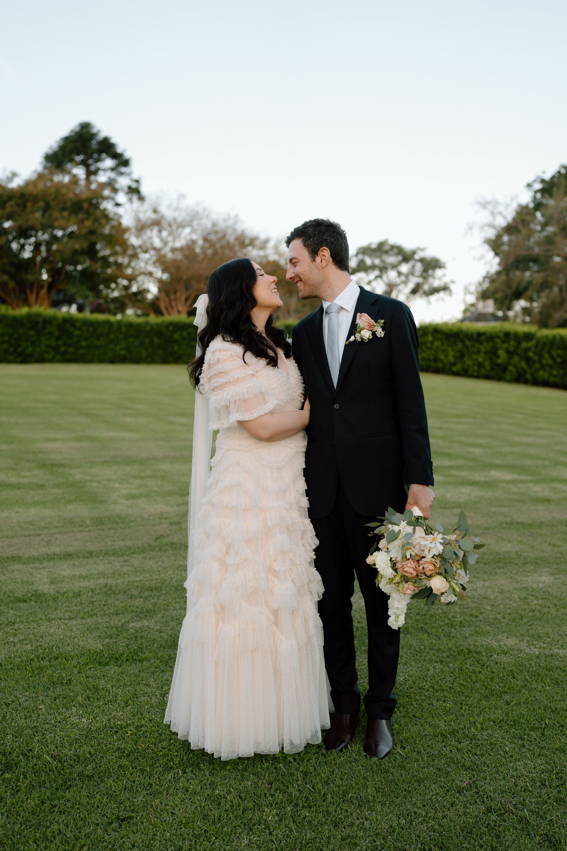 Bride and groom smile at each other on a grassy lawn. Bride in ruffled dress, groom in suit, holding bouquet.