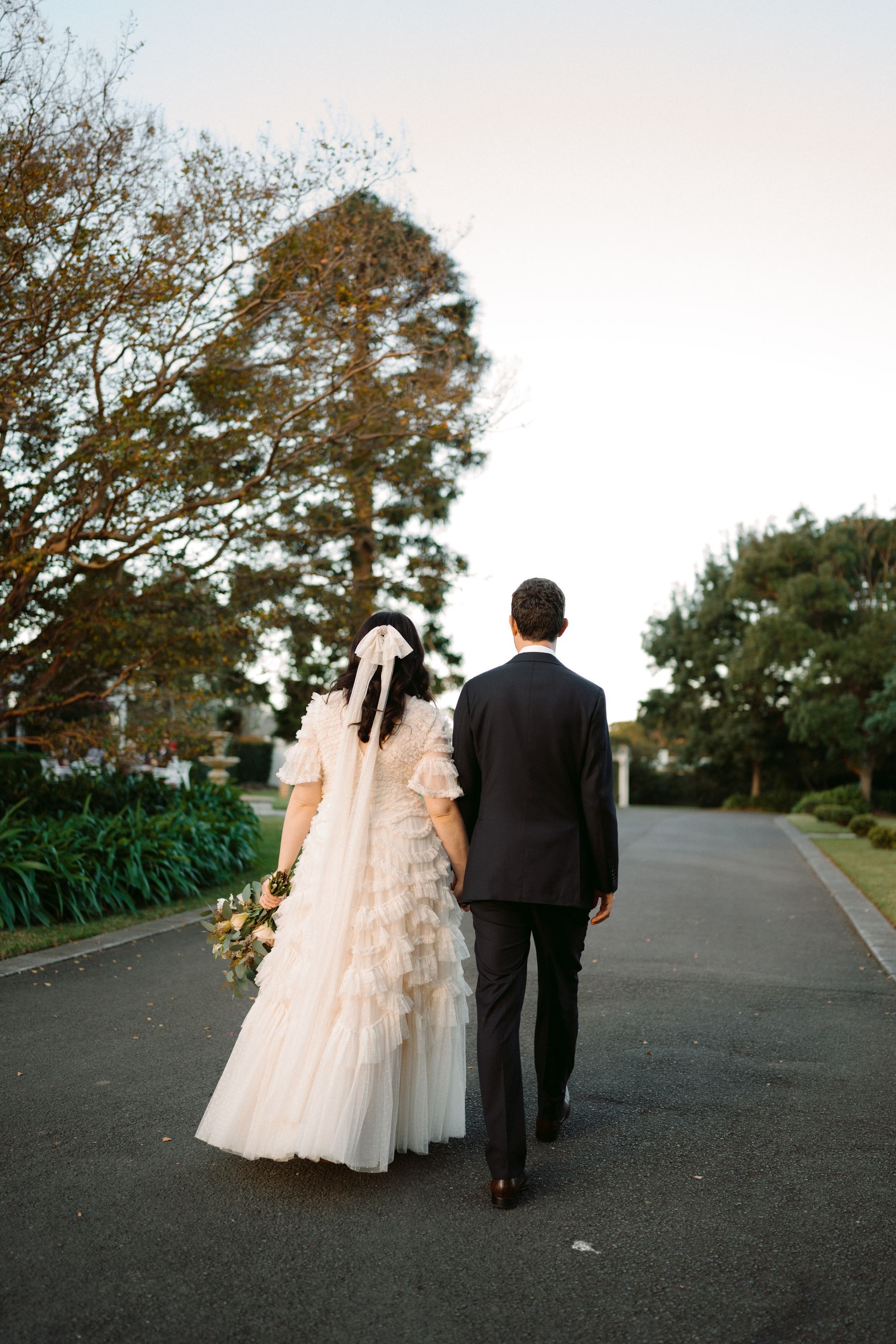 Newlyweds walk hand-in-hand down a tree-lined path; bride in a layered gown, groom in a suit.