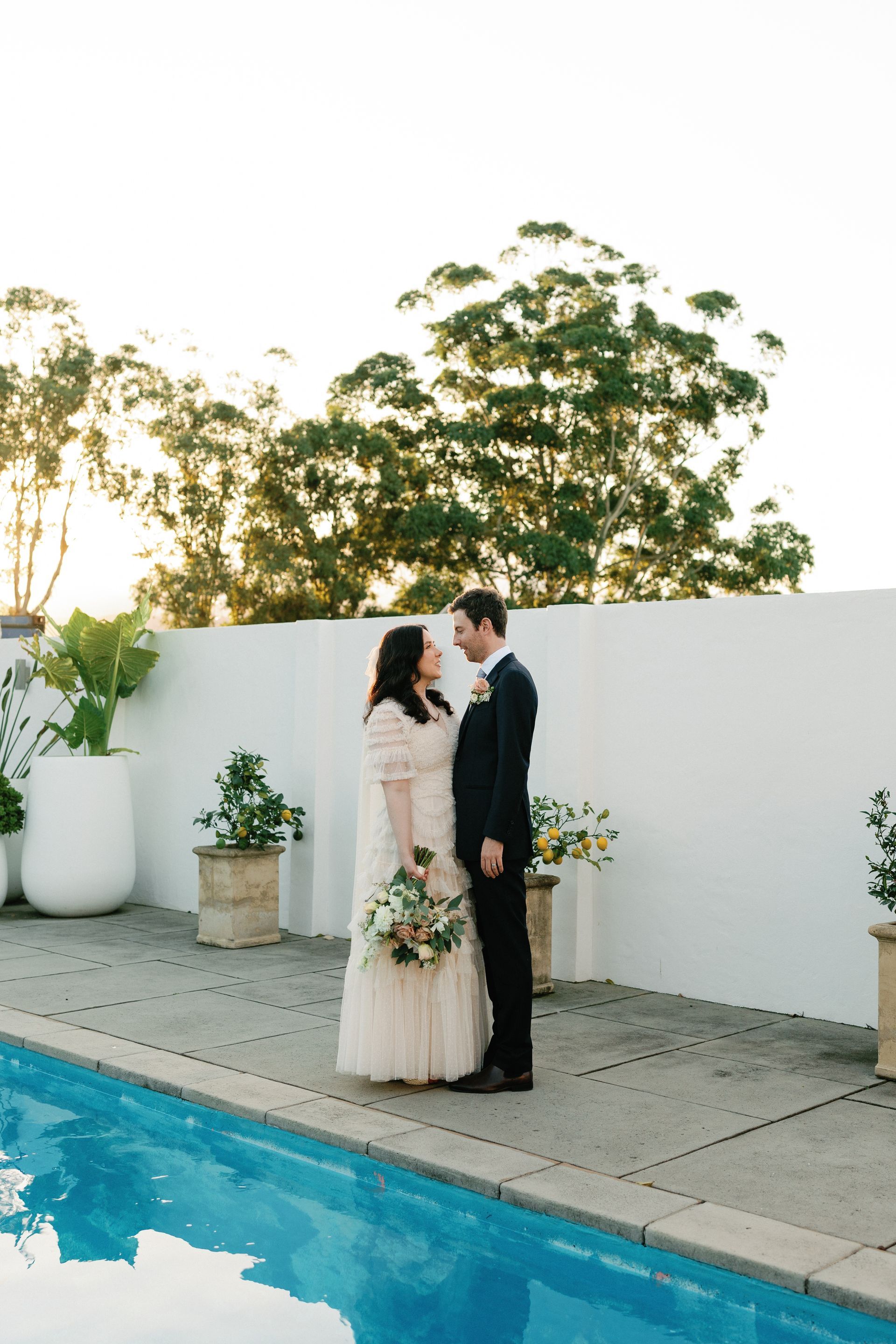Newlyweds in formal attire by a pool, looking at each other. White wall and trees in the background, golden light.