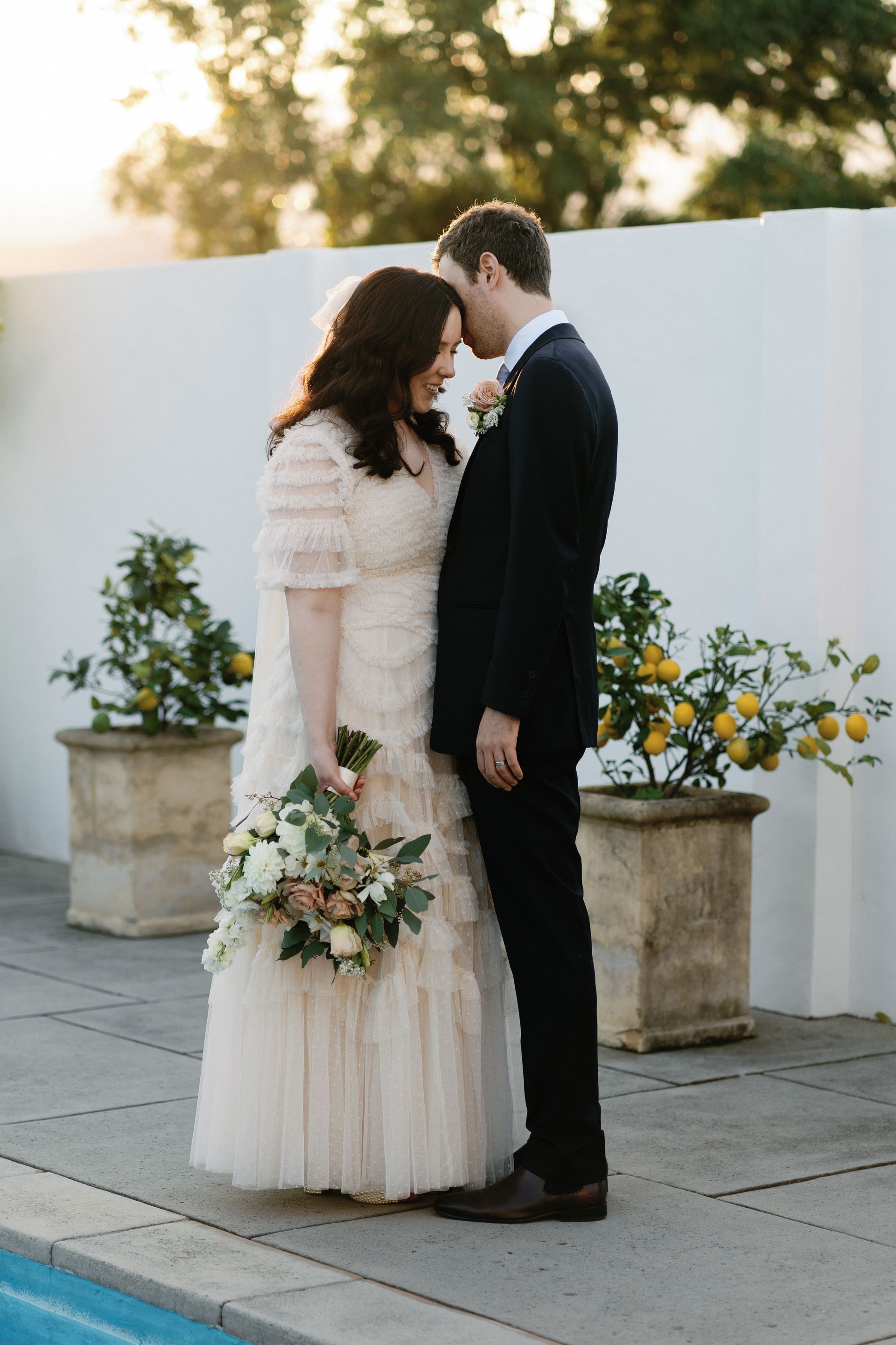 Bride and groom embrace outdoors near a pool, she wears a lace gown, he wears a navy suit.