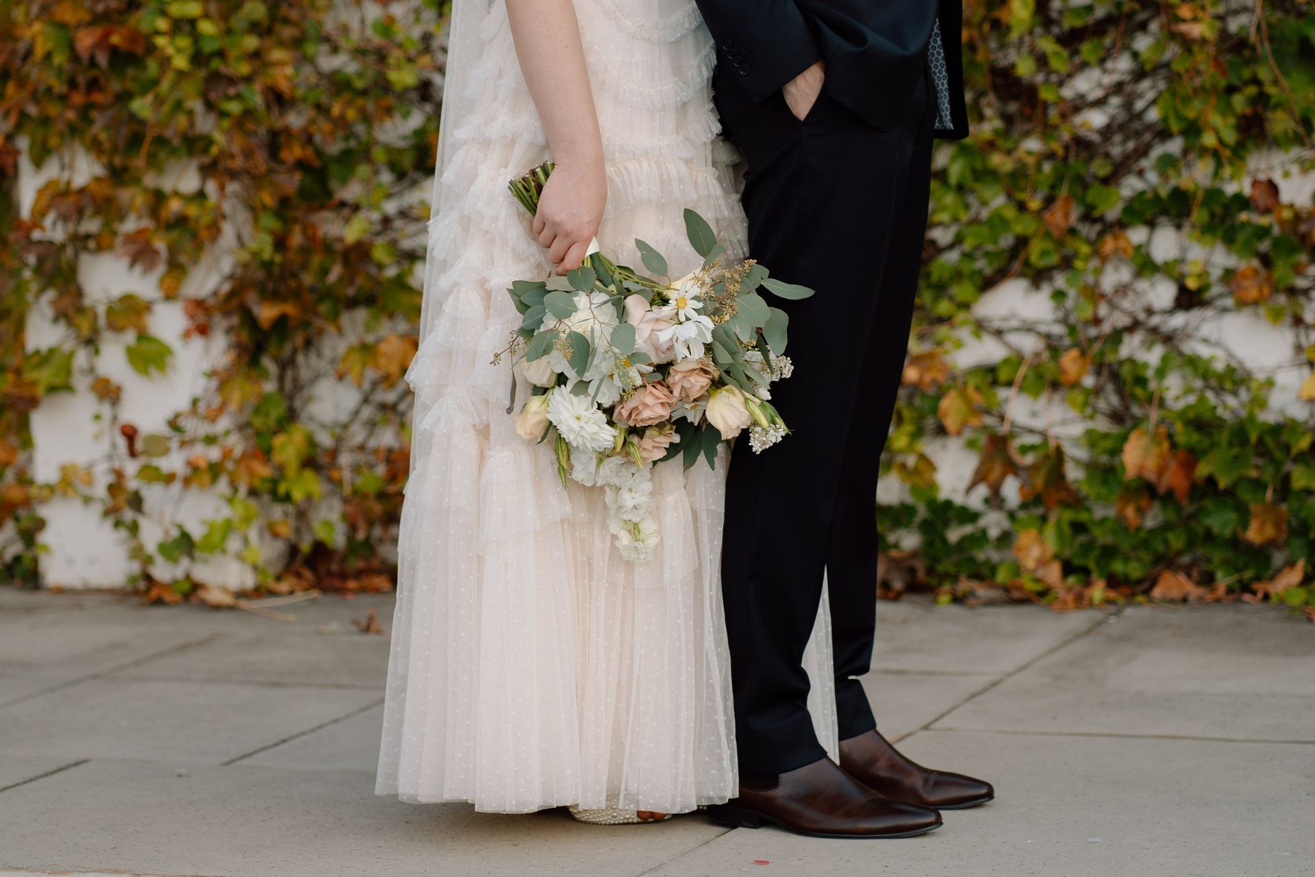 Bride in a white dress holds flowers, standing with a groom in front of a wall covered in vines.