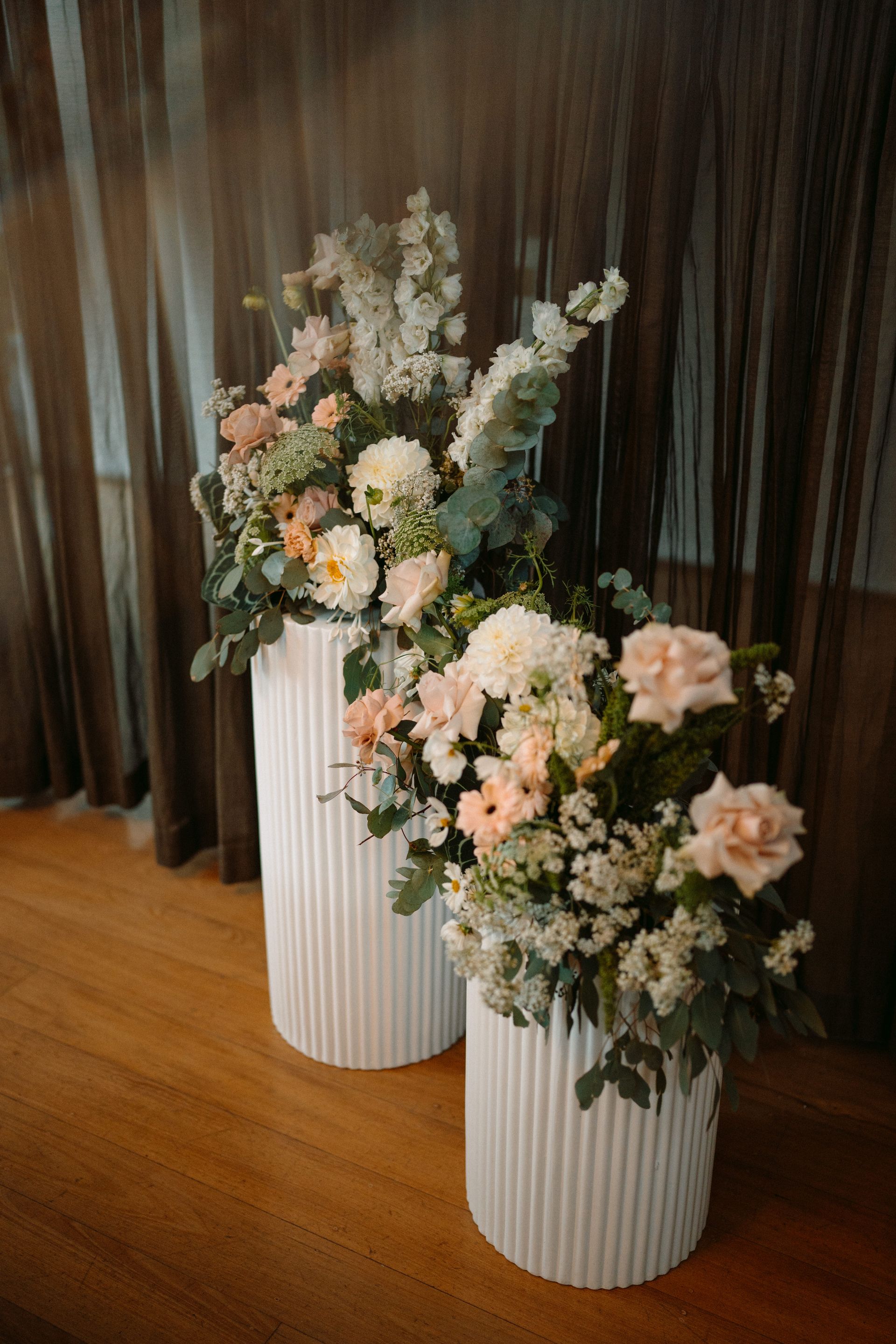 Two white fluted pedestals with pastel floral arrangements, set against dark curtains and a wooden floor.