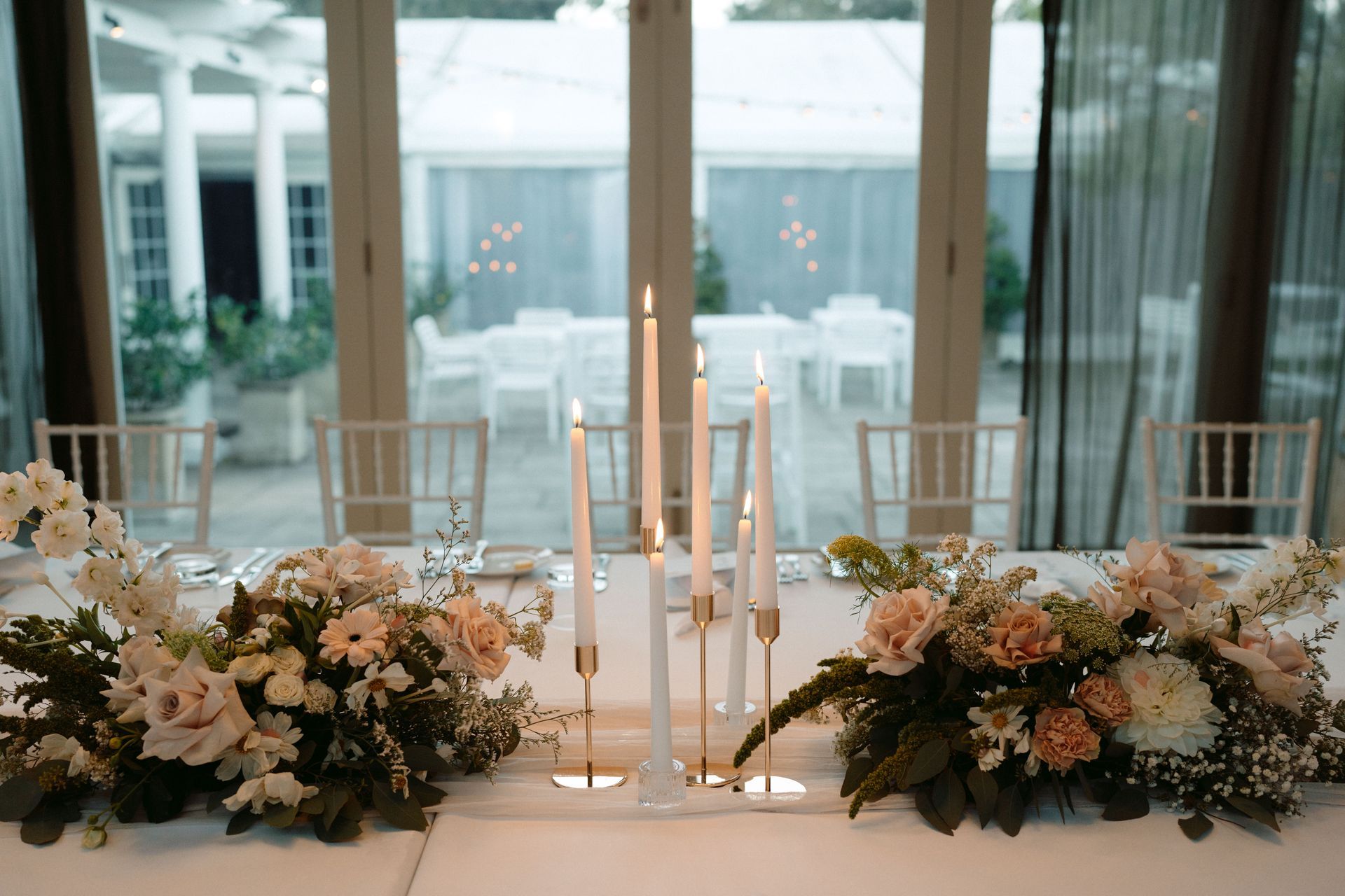 Wedding table with floral arrangements, candles, and outdoor setting visible through glass doors.