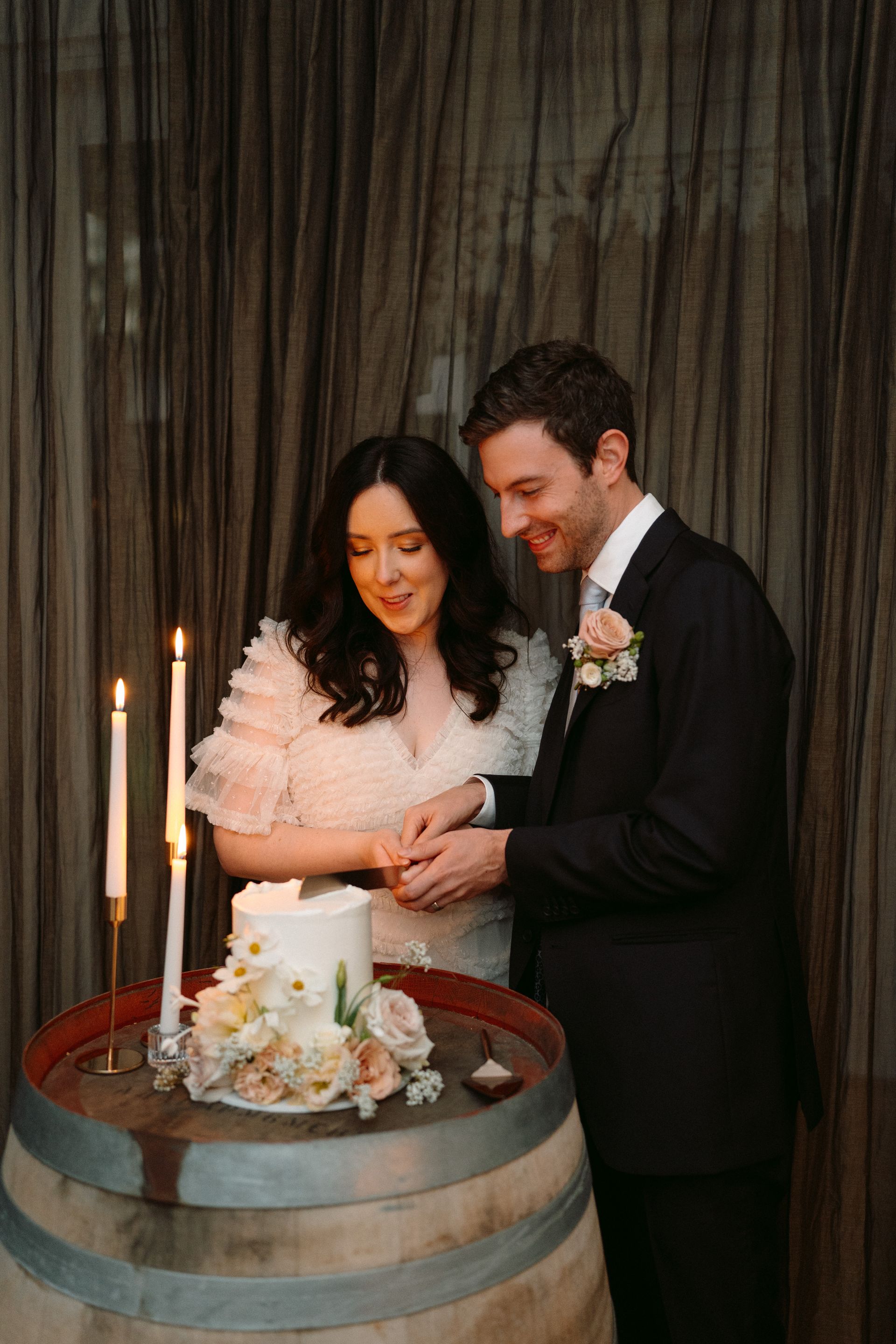 Couple cutting wedding cake on a barrel, surrounded by candles and a dark curtain backdrop.
