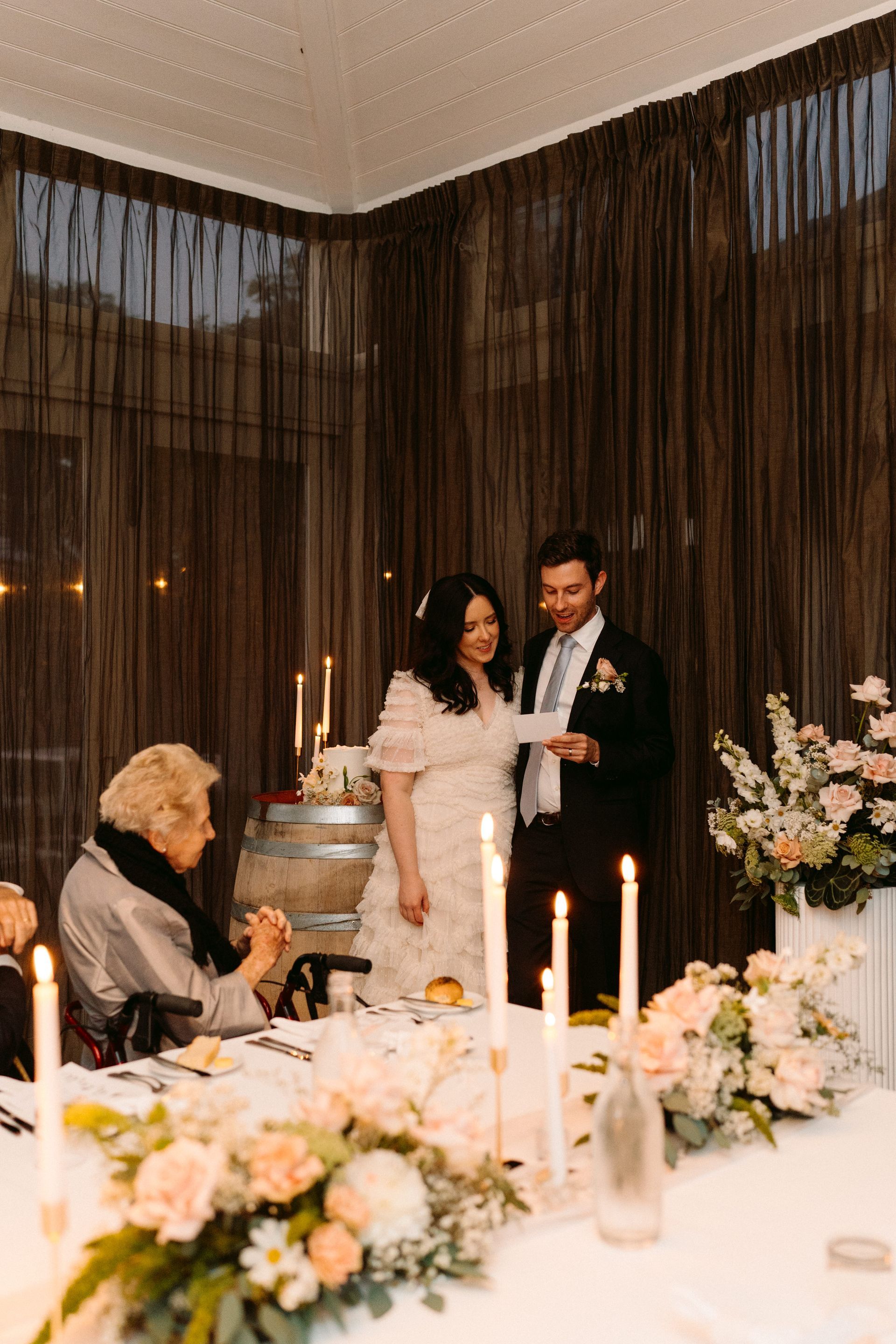 Bride and groom stand at a table reading, with guests. Floral centerpieces and candles decorate.
