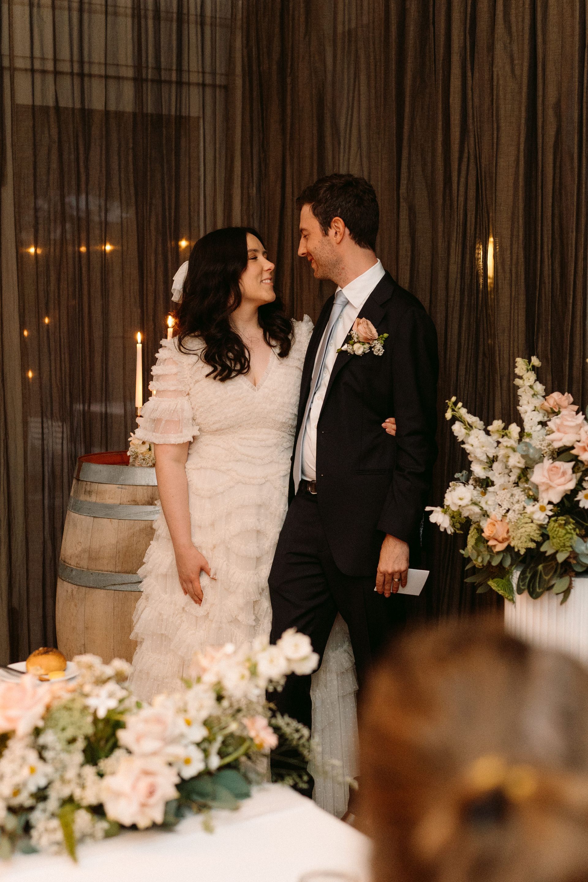 Newlyweds gaze at each other, smiling. Bride in a tiered dress, groom in a suit. Wedding reception setting, flowers.