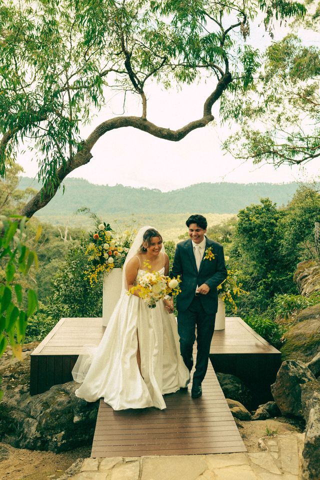 Bride and groom walking hand-in-hand on a wooden path, smiling, in a natural outdoor setting.