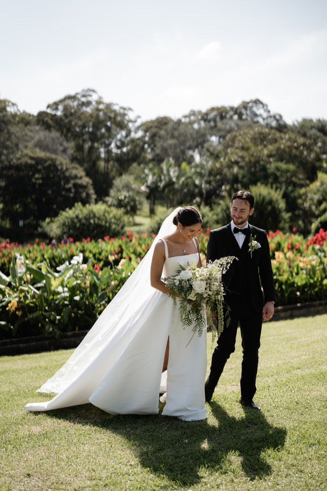 Bride and groom walking in a garden on wedding day. The bride wears a white gown and veil; the groom wears a black tuxedo.