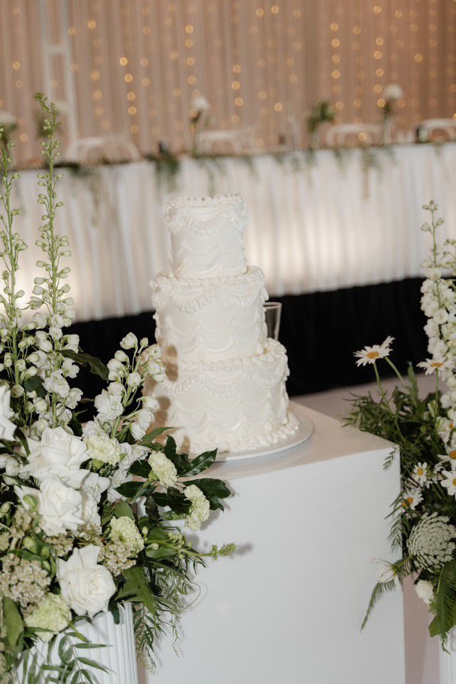 Three-tiered white wedding cake on a white pedestal, surrounded by white flowers and greenery.