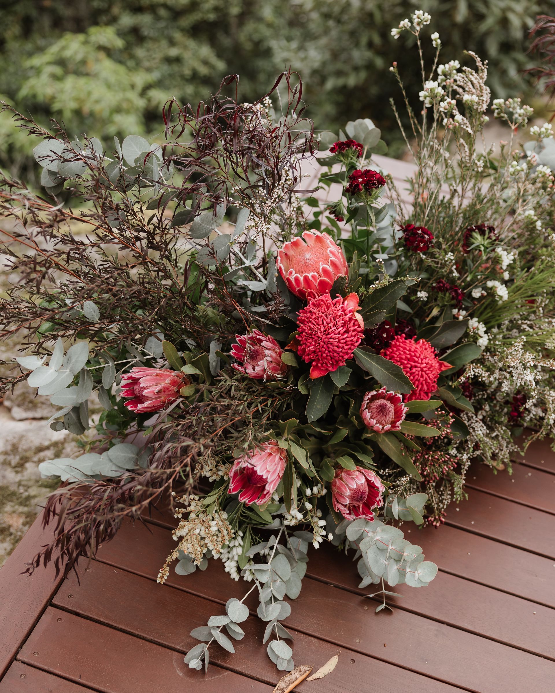 Floral arrangement with red protea, eucalyptus, and greenery on a wooden surface.