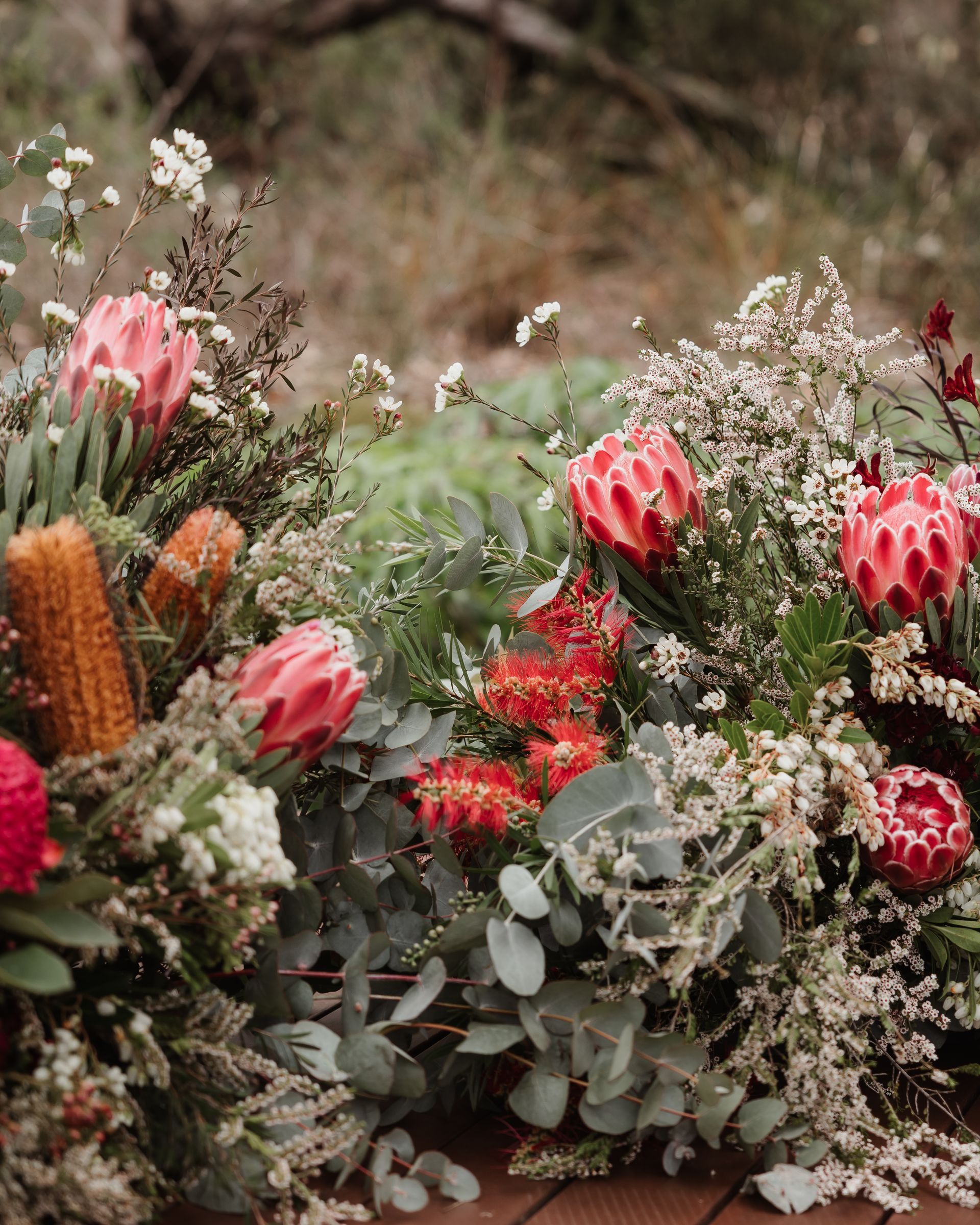 Floral arrangement with pink protea, orange banksia, and greenery against a blurry nature backdrop.