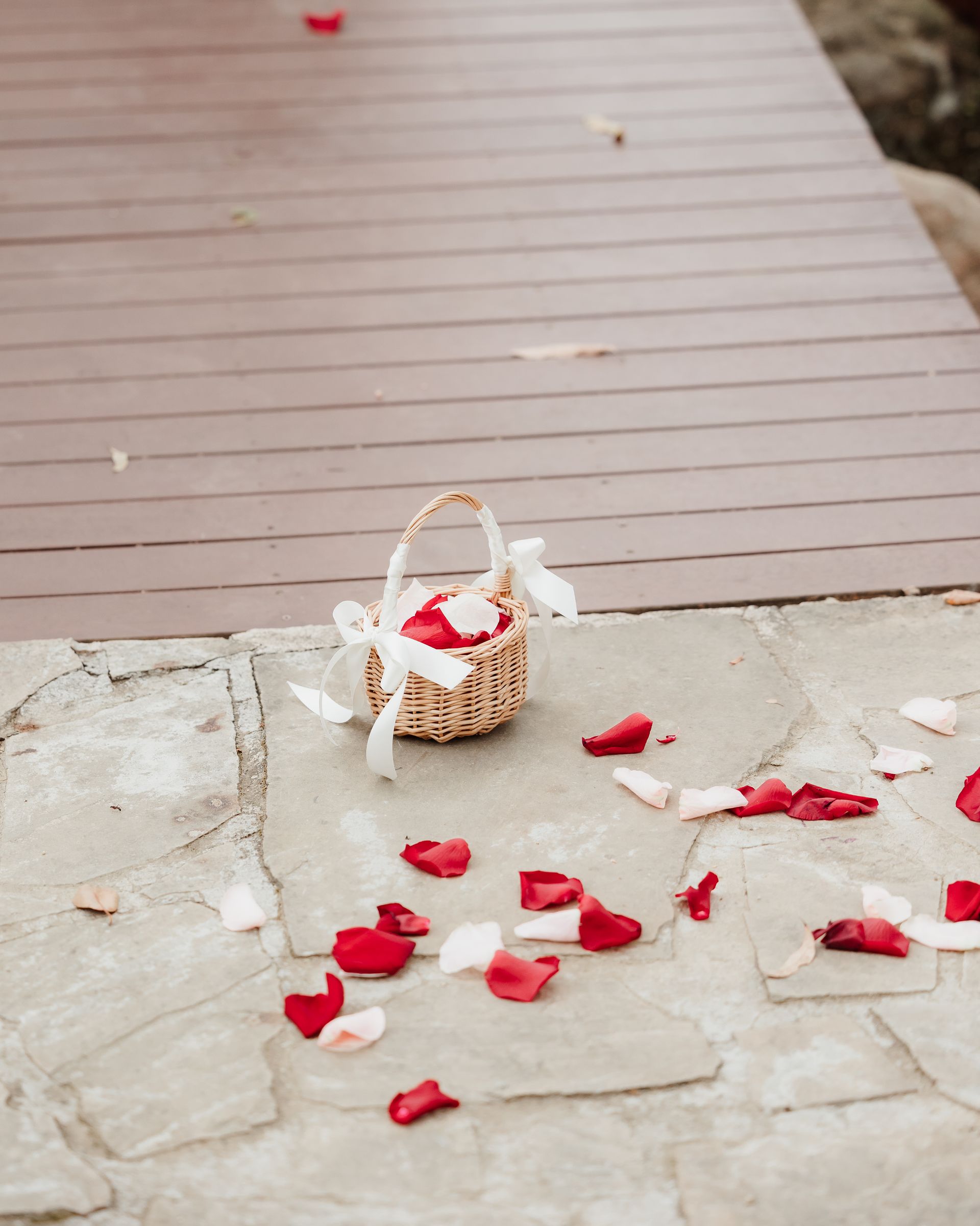 Wicker basket with white ribbon and rose petals scattered on stone and wood planks.