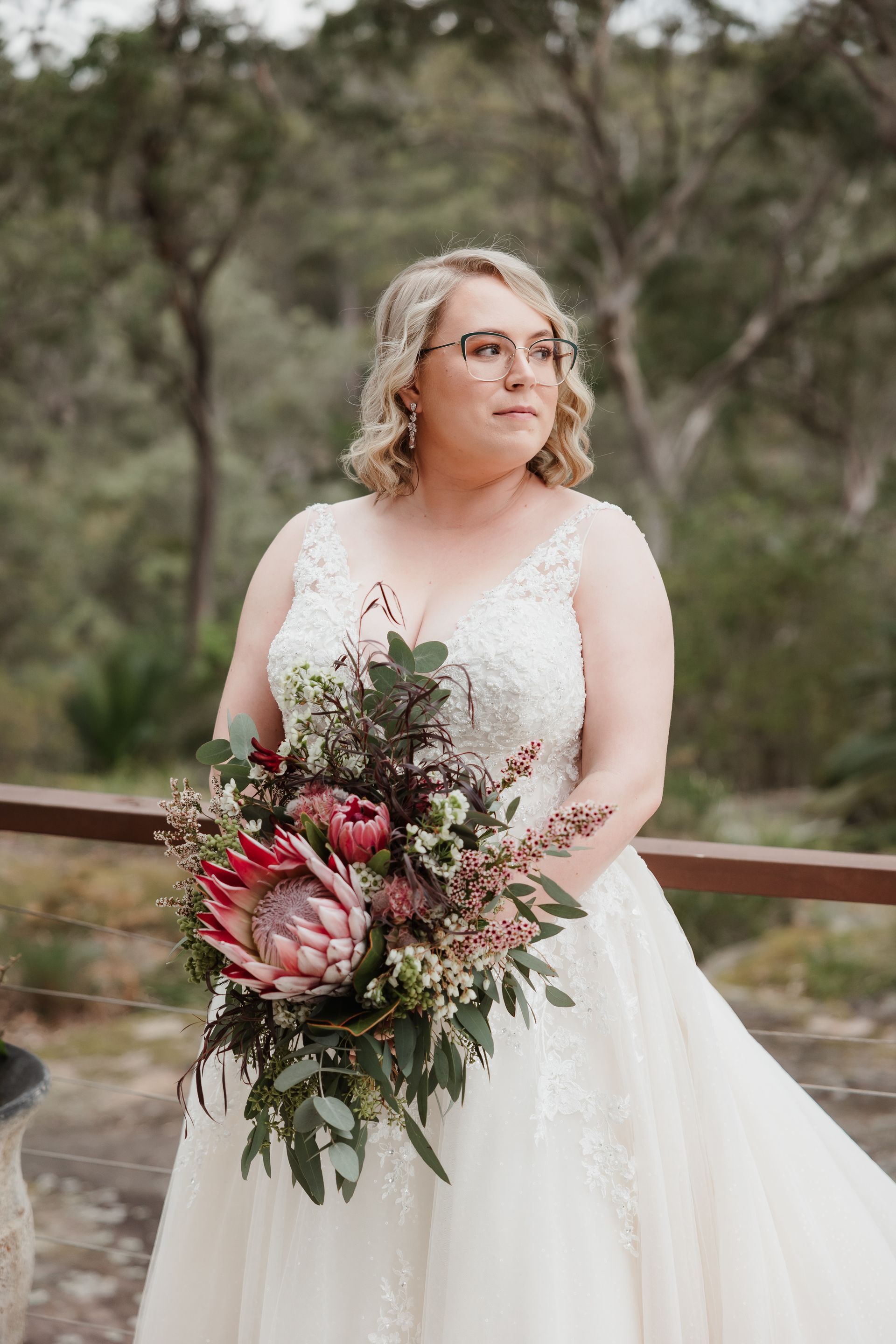 Bride in white dress, holding floral bouquet, stands on balcony looking to the side.