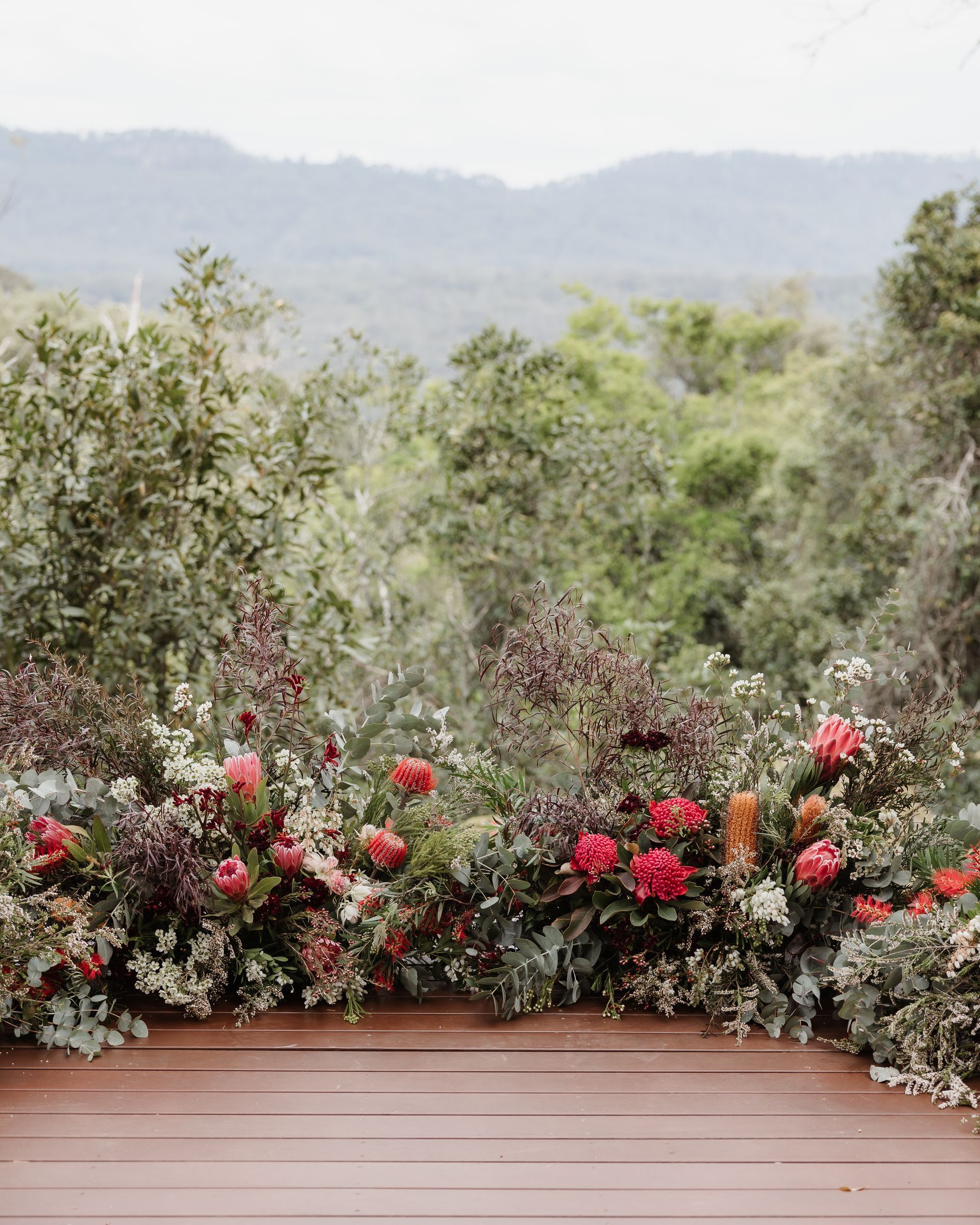 Wooden deck with a vibrant floral arrangement of red and burgundy flowers, overlooking a lush green forest.