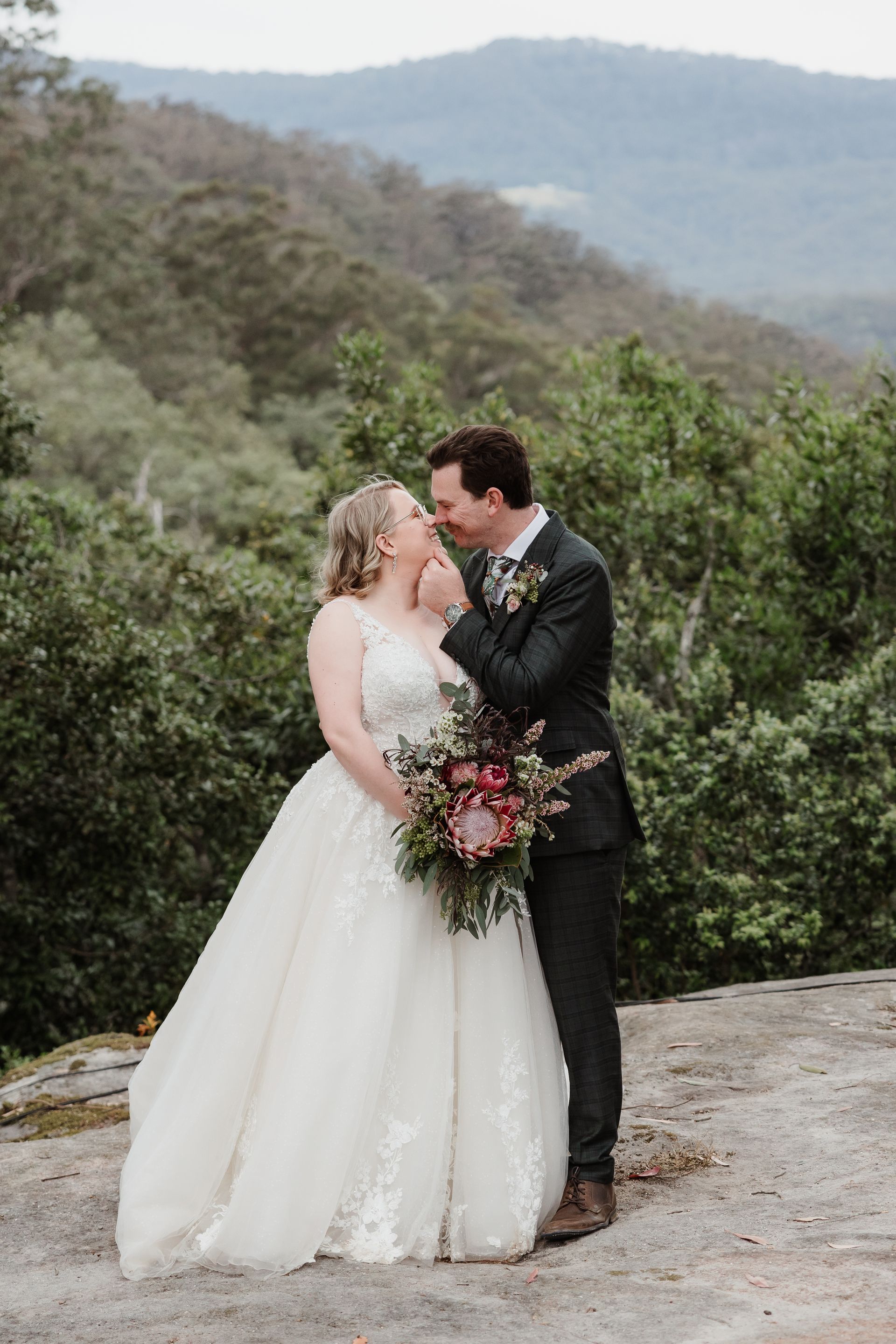 Bride and groom embrace outdoors on a rocky cliff, surrounded by green trees and a mountain backdrop.
