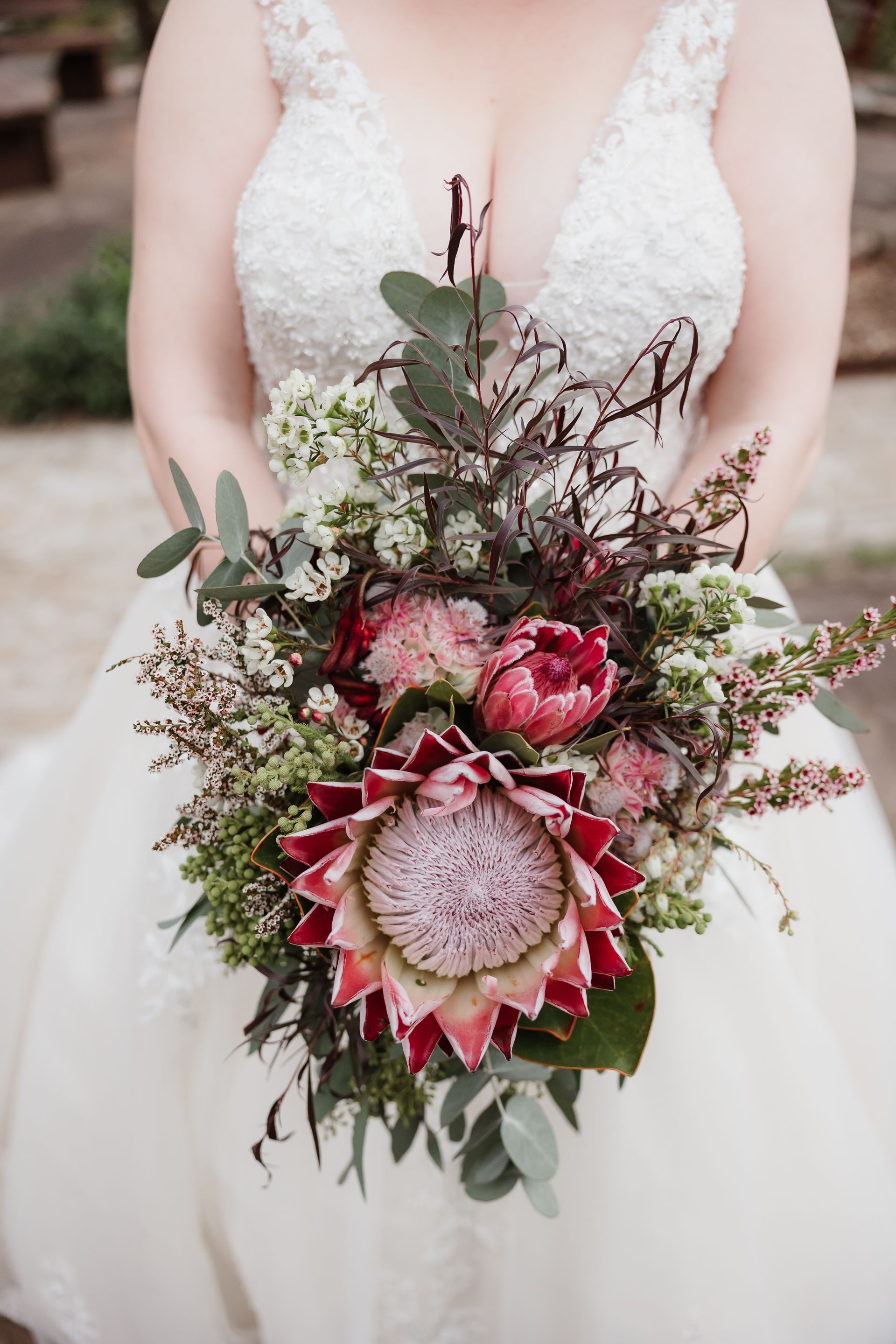 Bride holding a bouquet with a large pink protea flower and greenery, wearing a white lace wedding dress.